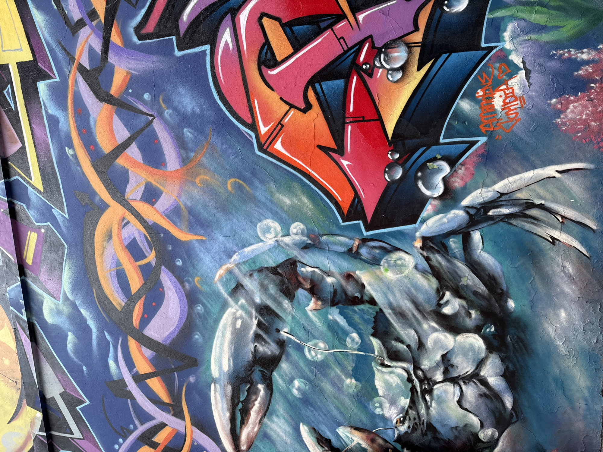 The image depicts a close-up of a mural, possibly located in Buenos Aires, Argentina given the prompt’s context. The mural features an illustration of a crab with large claws surrounded by air bubbles. Above the crab is a graffiti-style design with red, orange, and blue colors. To the left are abstract orange and purple swirls. The background is a blend of blue tones suggesting an underwater environment. A section of orange lettering is also visible at the top right of the crab. The surface is textured with cracks and weathering, indicating that the mural is outdoors and exposed to the elements.