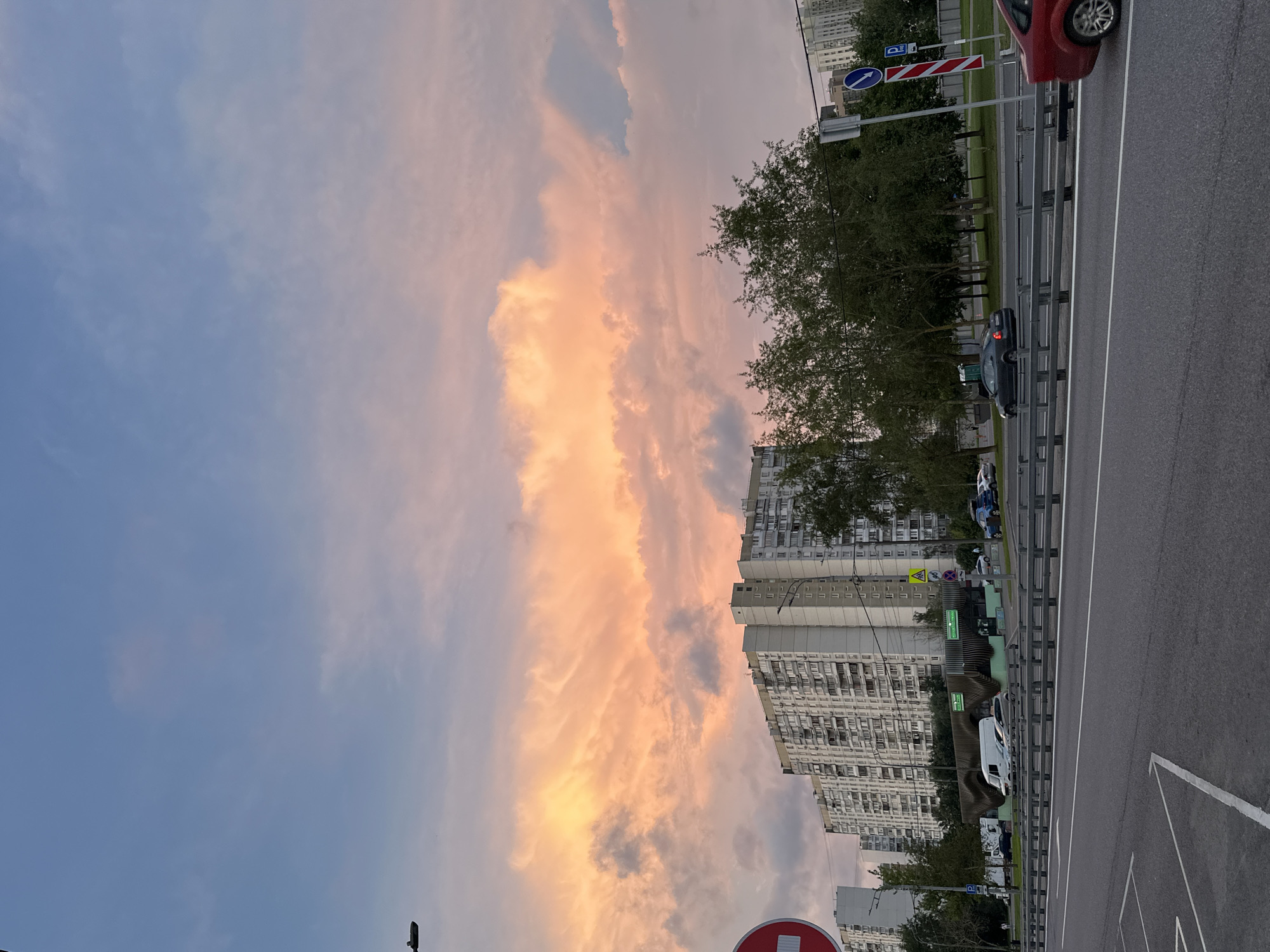 The image captures a street scene in Moscow, Russia, during what appears to be sunset. The sky is dominated by dramatic clouds tinged with orange and pink hues, contrasting with the pale blue sky above. In the background, a long, multi-story apartment building with numerous windows is visible. In front of the building, a line of trees forms a green band. A multi-lane road occupies the foreground, with cars driving in both directions. A red car is visible on the right side of the frame, and a dark sedan is in motion in the middle lane. Traffic signs, including a "No Entry" sign and a parking sign, are present along the road. A white van is parked on the side. The overall impression is an urban environment with a striking natural sky phenomenon.