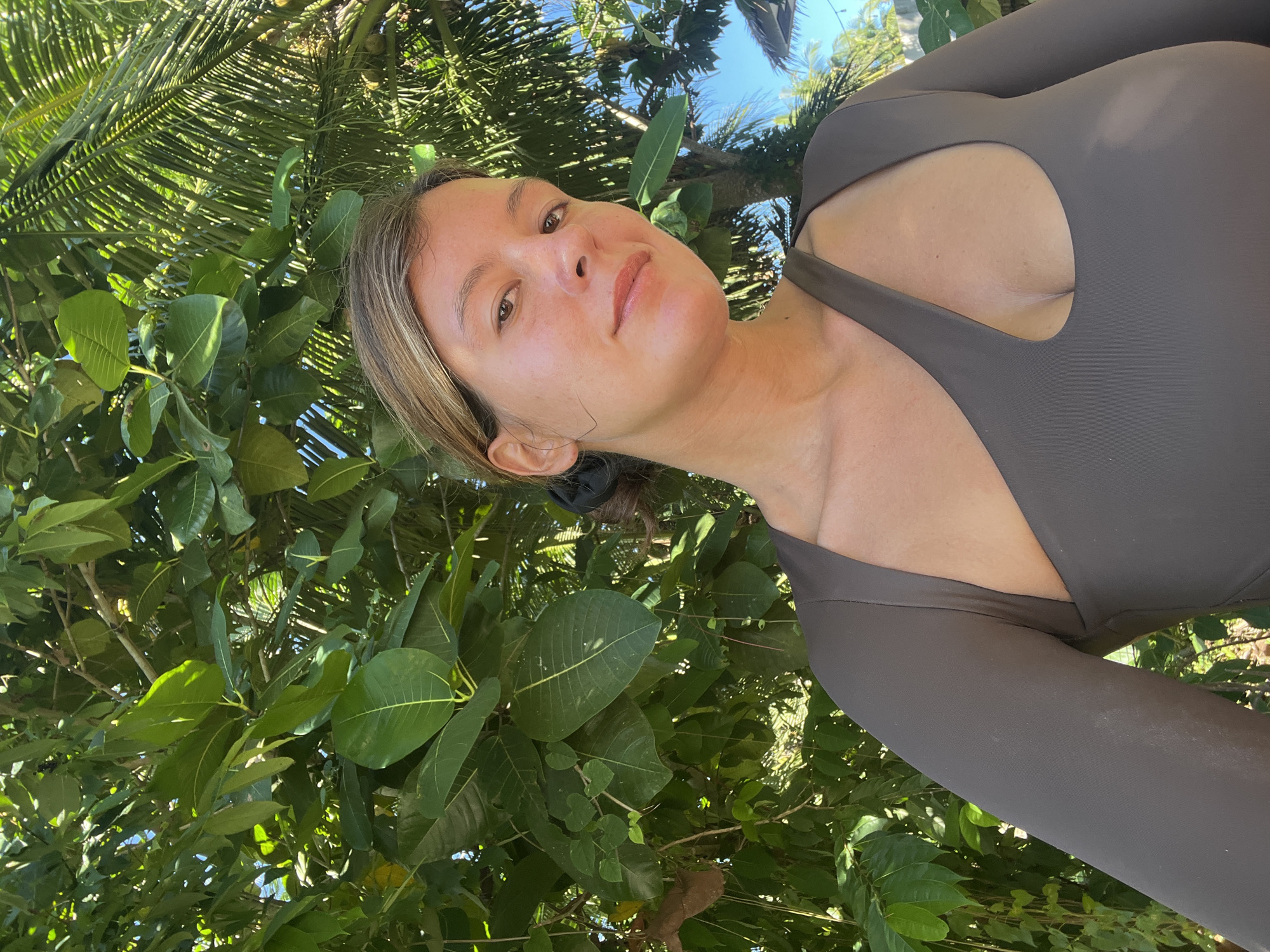 A woman with light skin and brown hair is taking a selfie outdoors. She is wearing a brown, one-sleeved swimsuit with a cutout over her chest. She has her hair pulled back in a bun and is looking towards the camera. The background is filled with lush green foliage and palm trees, with a clear blue sky visible. The image appears to have been taken during the daytime in a tropical location, consistent with General Luna, Philippines.