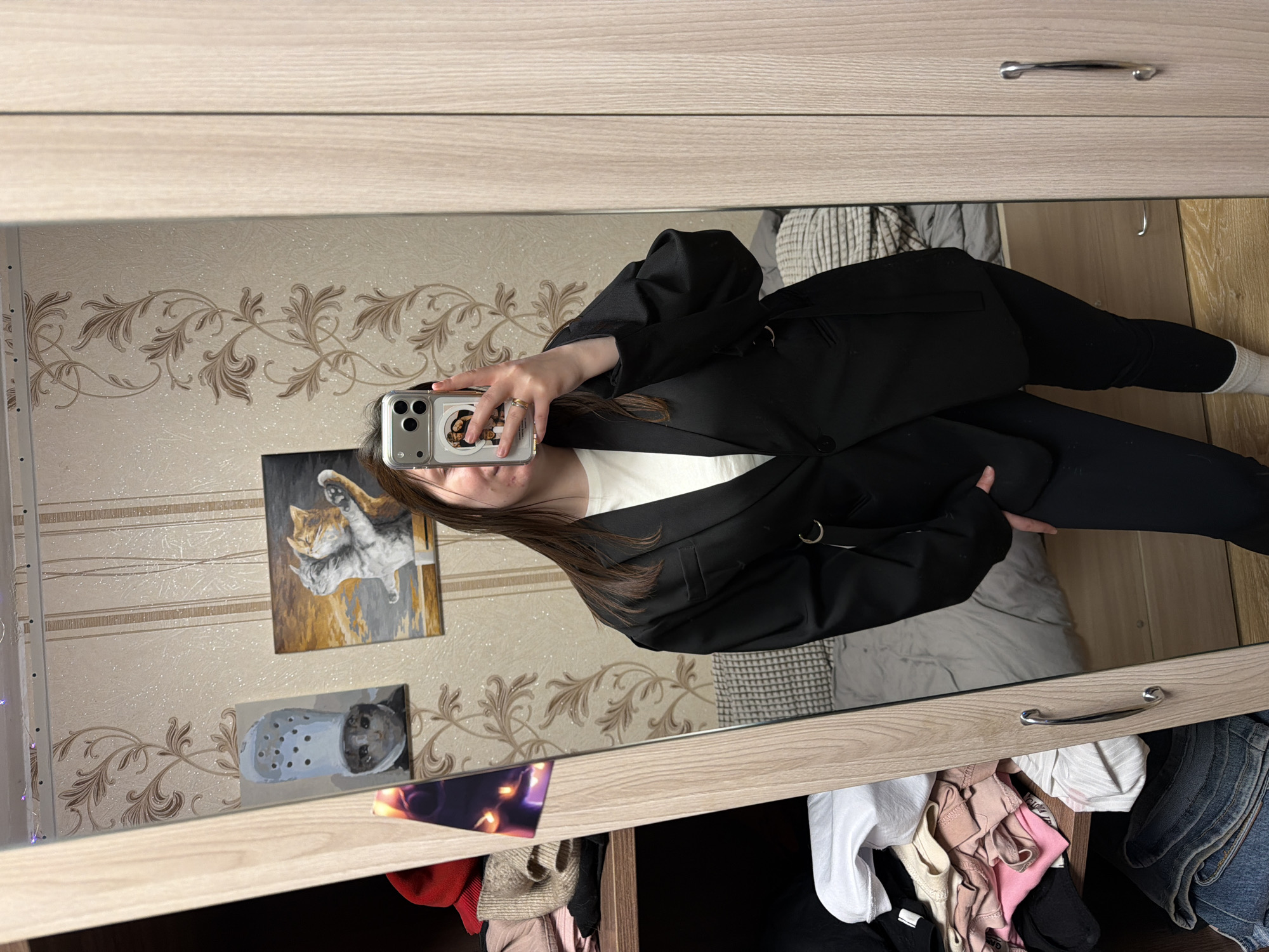 A mirror selfie shows a woman indoors, likely in an apartment in Chaikovsky, Russia. She wears a black blazer, black leggings, and a white shirt. She holds a phone with a sticker on the back, obscuring her face. The background shows floral wallpaper, a painting of cats, and another painting of a cat inside a Croc shoe. A drawer beneath the mirror is open, filled with clothes.