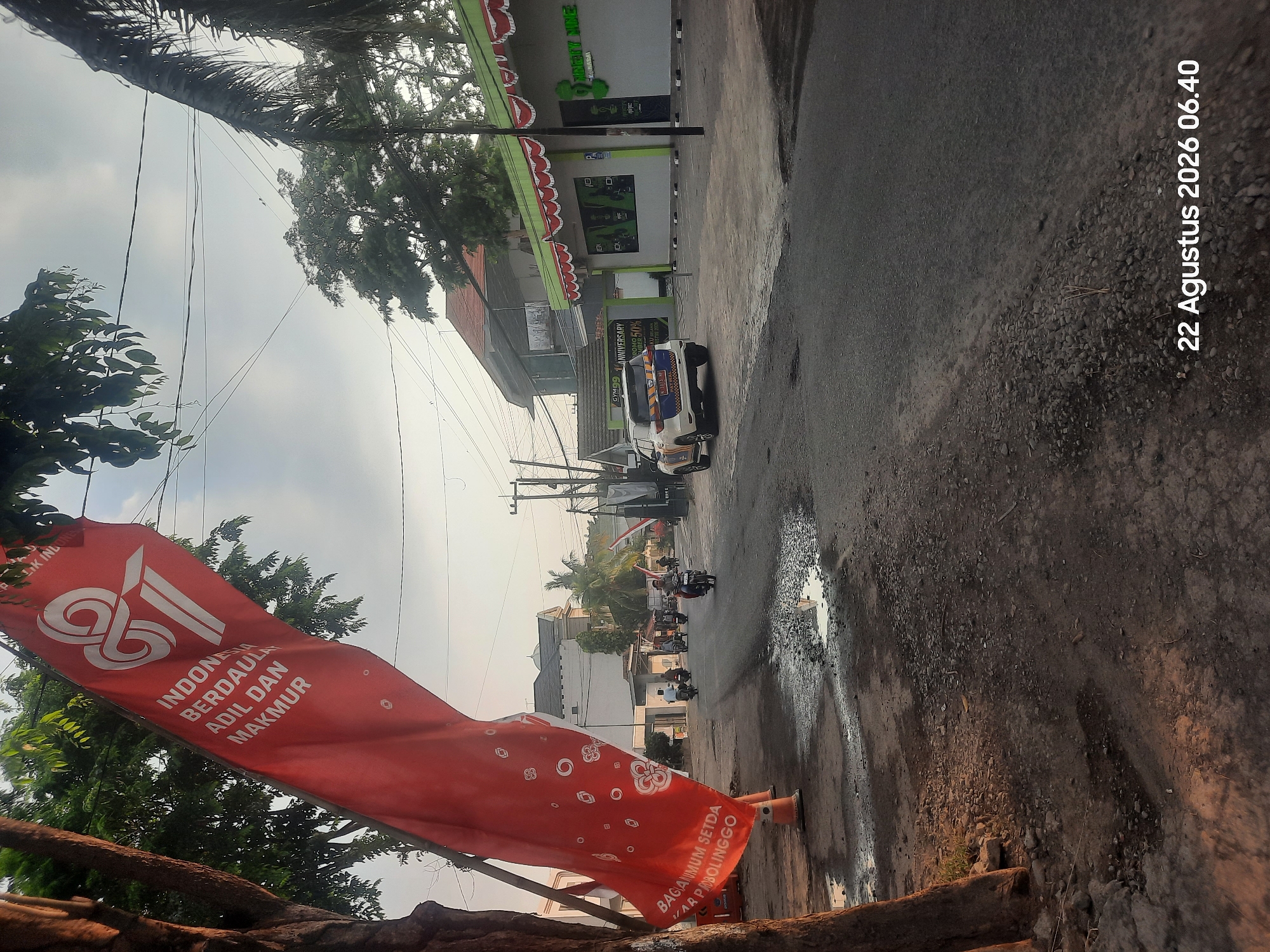 The image captures a street scene in Pajarakan Kulon, Indonesia, with a red banner prominently displayed in the foreground. The banner, emblazoned with white text and a stylized graphic, reads "INDONESIA BERDAULAT ADIL DAN MAKMUR" and "BAGIANUM SETDA KAB PROBOLINGGO". Behind the banner, a road with some puddles is visible, with several motorcycles and a police car driving on it. Buildings line the street, including a structure with green and white awnings. Trees and power lines are present throughout the scene, and the sky is overcast. The date "22 Agustus 2026 06.40" is superimposed on the bottom right corner of the image.