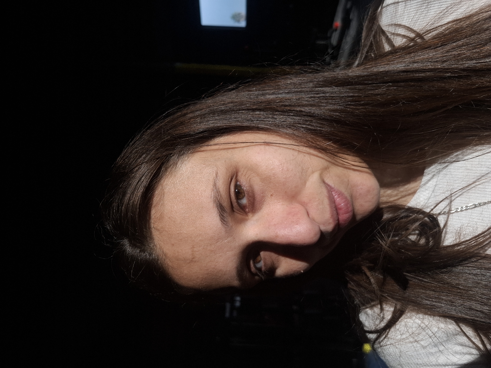 A medium shot captures a woman's face and upper chest against a dark background. She has long, dark brown hair that falls around her shoulders. Sunlight illuminates the left side of her face, casting a shadow over the right. Her skin appears light-toned with some texture visible. Her eyes are brown and looking slightly off-camera. She is wearing a white, textured shirt, and a silver chain necklace is visible. The background is mostly dark, with a dimly lit square or rectangular object visible in the upper right quadrant, possibly a screen, with a faint image of a tree or plant on it. There are also some indistinct shapes and lights in the darker areas of the background. The setting appears to be indoors, possibly in a studio or a dimly lit room, in the city of Wilde, Argentina.