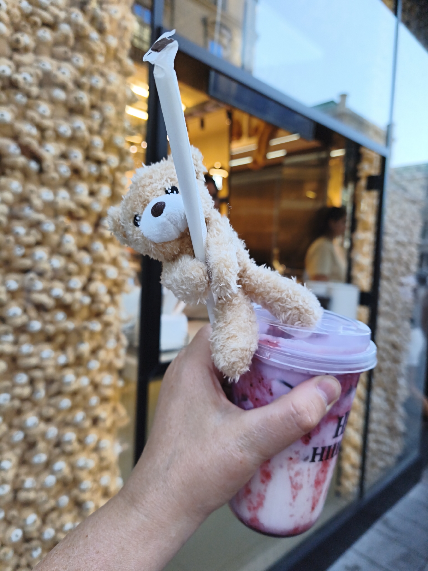 A person's hand holds a clear plastic cup filled with a pinkish-white beverage and streaks of red fruit. A small, light brown, fuzzy teddy bear is attached to a white straw, with its arms wrapped around the straw. The straw is inserted into the beverage. The cup is sitting on a counter, with a blurry background that appears to be a shop window. Behind the window, a person is visible, and the exterior of a building with an ornate facade is also present. The overall scene suggests a casual outing or a stop at a cafe or food establishment. The location is identified as Vienna, Austria.