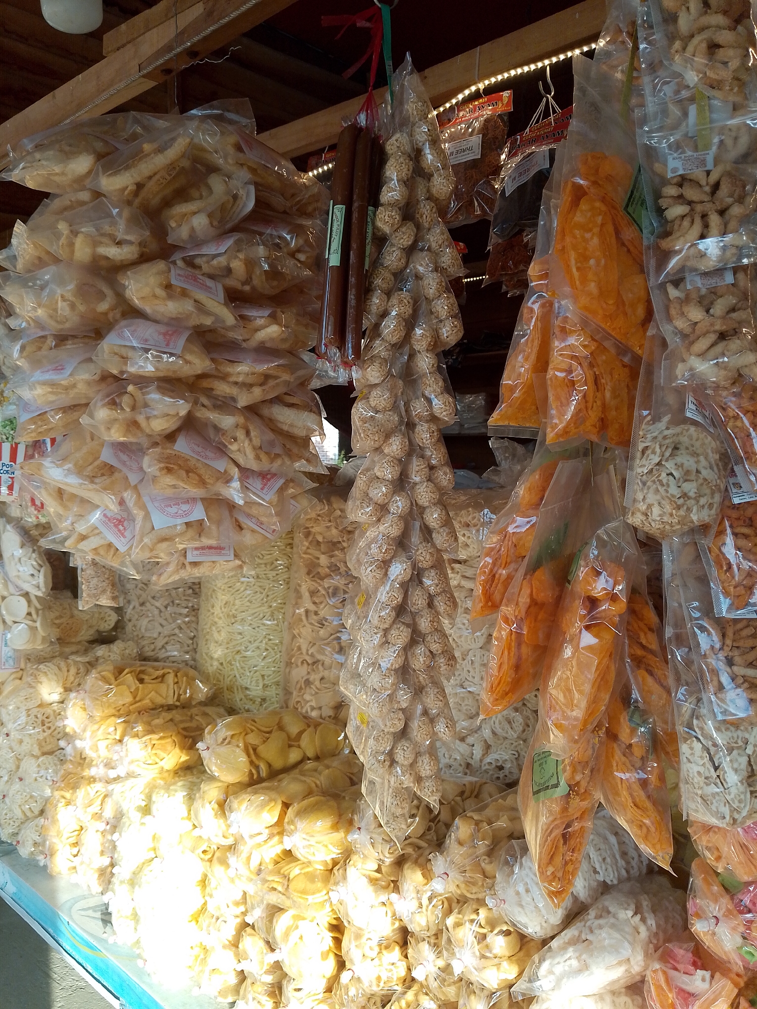 This image depicts a market stall in Sawang, Indonesia, overflowing with a variety of packaged snacks. The stall is densely packed with goods, primarily displayed in transparent plastic bags hanging from wooden beams or stacked on shelves. The snacks themselves are diverse in shape, size, and color, ranging from small, round, puffed items to flat, crisp slices and long, noodle-like strands. Many of the packages have labels, though the text is largely illegible. The overall impression is one of abundance and variety, showcasing a typical market scene focused on the sale of dried and packaged food items.