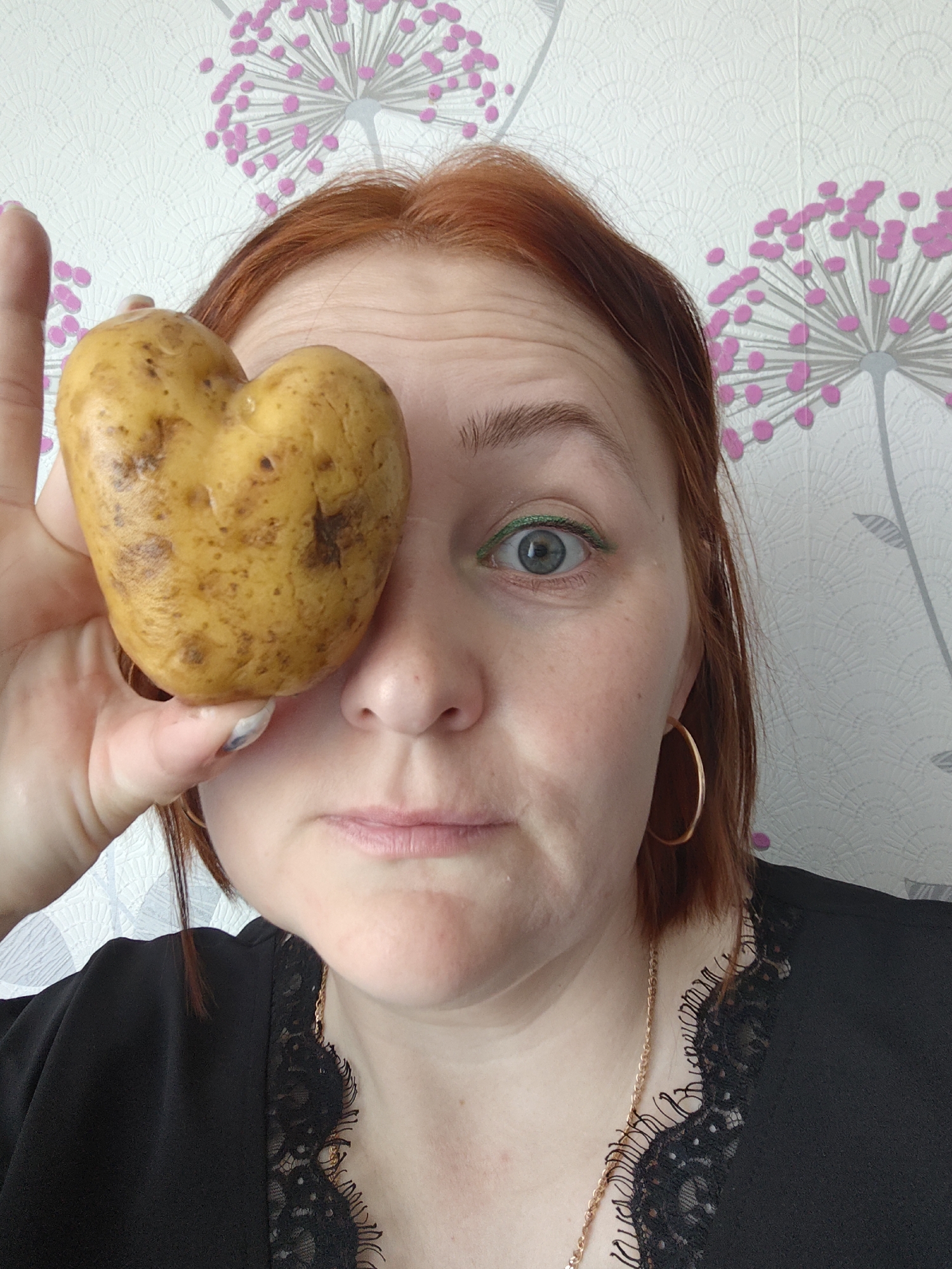 A woman with red hair and green eyeliner is holding a heart-shaped potato in front of her eye. She is wearing a black top with lace trim and gold jewelry. The background is a white wall with a floral pattern. The location is Markovskii, Russia.