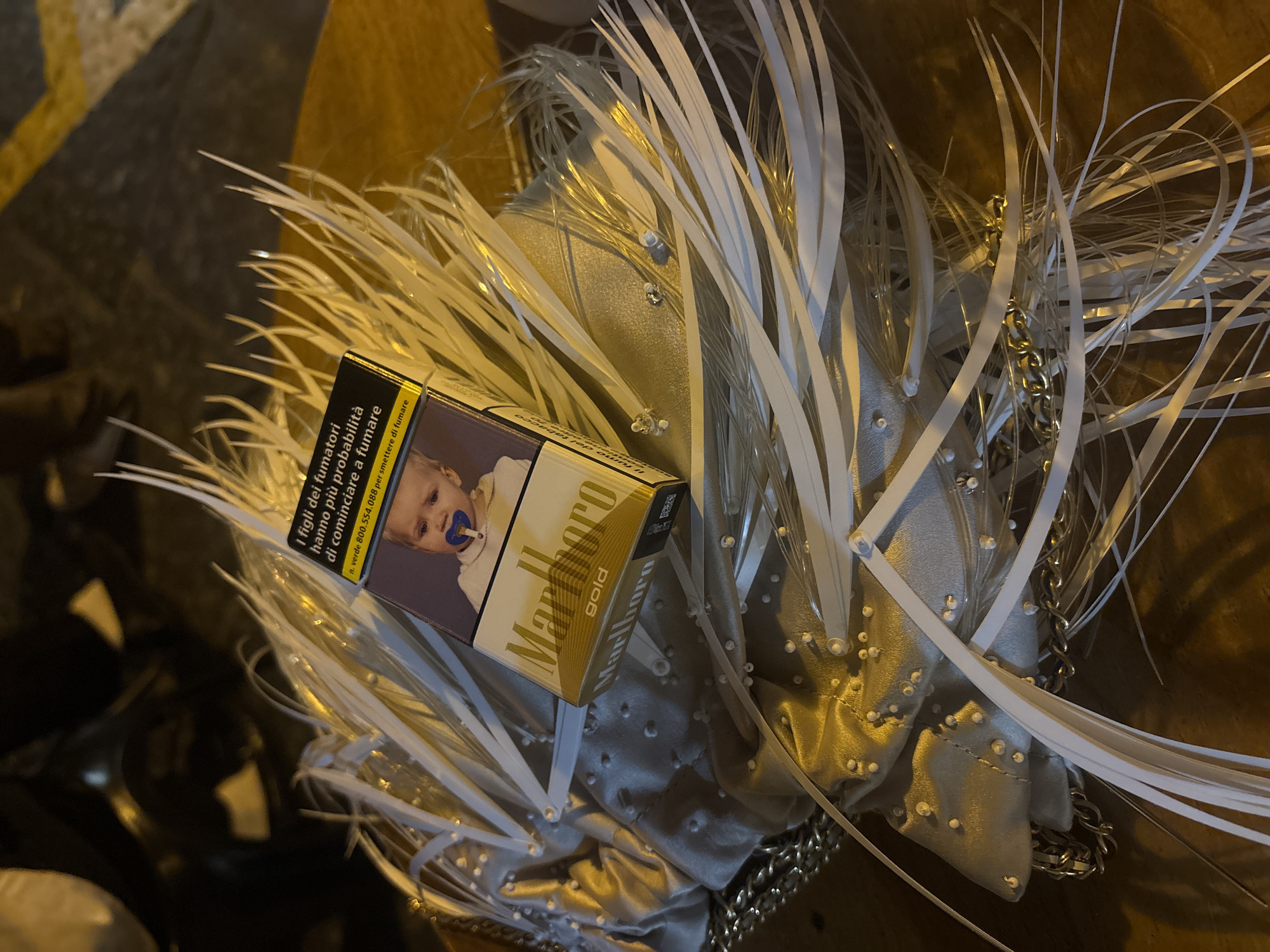 The scene shows a pack of Marlboro Gold cigarettes placed on top of a silver-colored decorative item, possibly a hat or handbag, adorned with white plastic strips and small silver studs. The cigarette pack features a warning label with an image of a baby and text in Italian. The background is blurred, suggesting an outdoor setting with a brown surface, possibly a table or the ground. The lighting is dim, and the overall composition is somewhat chaotic. The image may have been taken in Lipari, Italy.