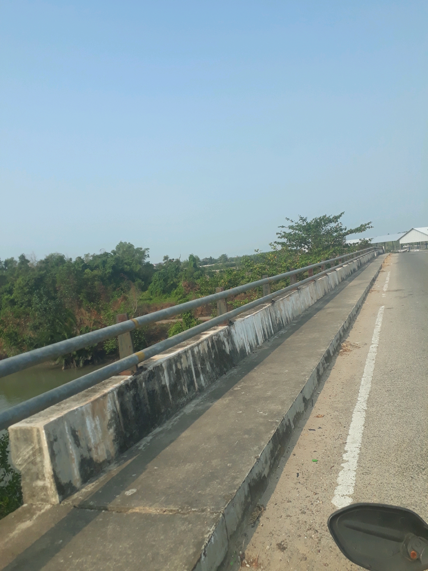 A clear, pale blue sky dominates the upper portion of the image. Below the sky is a landscape featuring lush green trees and vegetation on either side of a bridge. The bridge itself is made of concrete with metal railings. A paved road with a white line marking runs along the right side of the bridge. In the foreground on the far right, a portion of a black rearview mirror, likely from a motorcycle, is visible. The scene appears to be outdoors, possibly in a rural or semi-rural area, given the natural vegetation and the bridge over what appears to be a body of water. This image was captured on a bridge in Pangkalpinang, Indonesia.