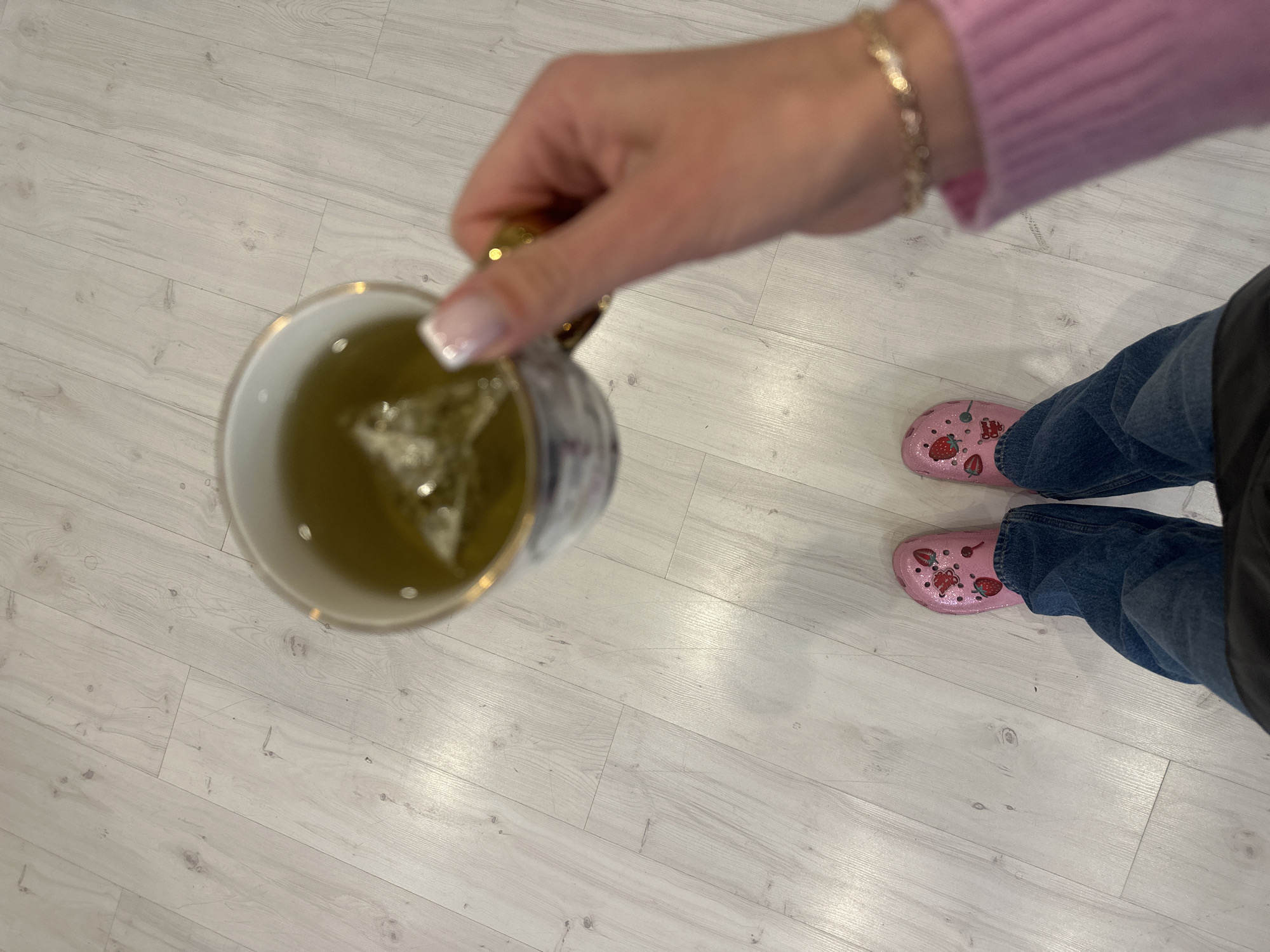 The photograph, taken from a high angle, presents a view of a person holding a mug with a tea bag inside. The liquid in the mug is a light yellow color. The person is wearing a pink long-sleeved shirt and blue jeans. On their feet are pink Crocs with strawberry charms. The background shows a light-colored, wooden floor. There is no indication of the city of Riga, Latvia.