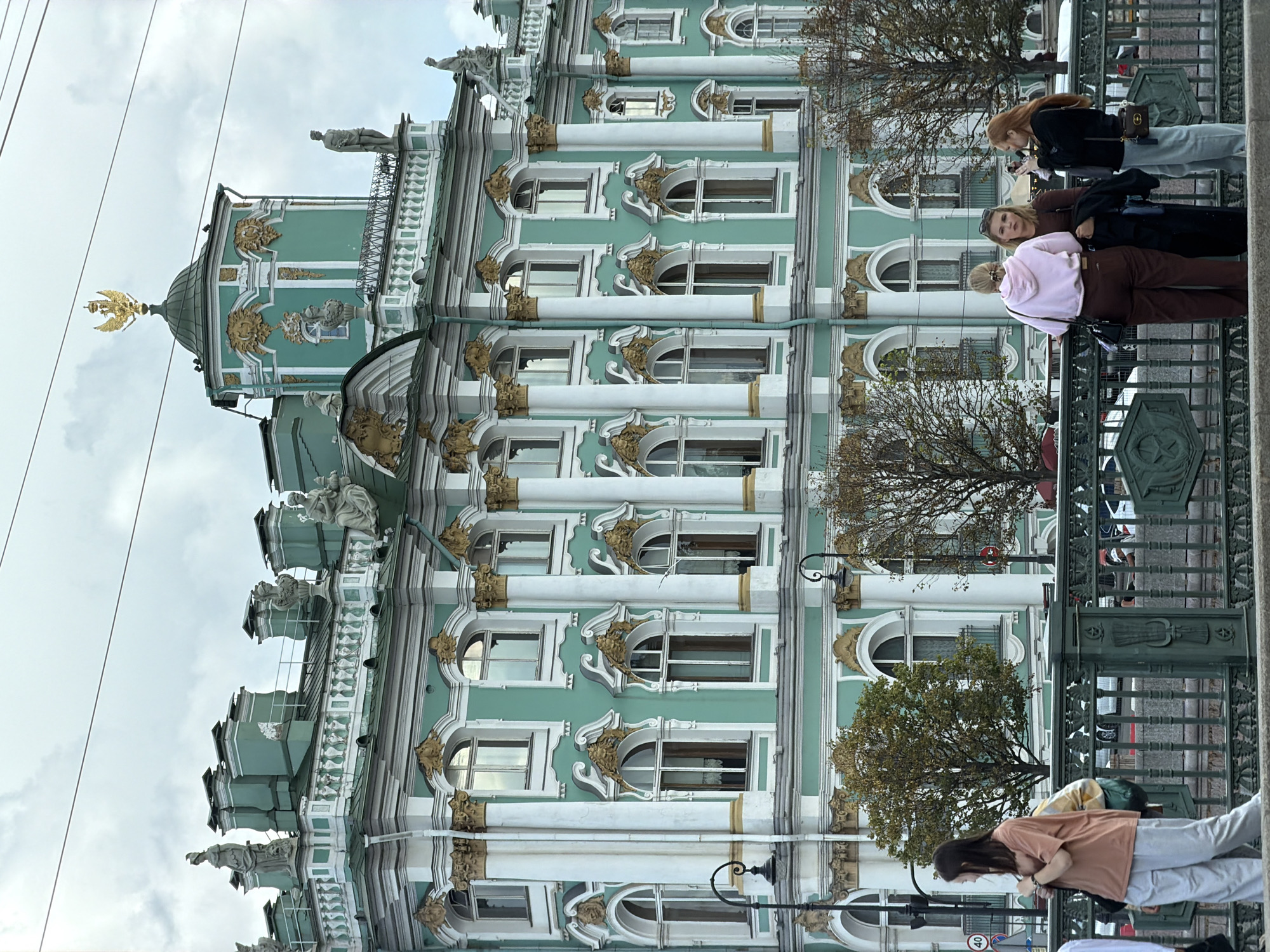 A group of four young women stand on a pedestrian bridge in front of the iconic green and white facade of the Winter Palace in Saint Petersburg, Russia. The palace, a masterpiece of Baroque architecture, is adorned with ornate gold detailing and classical sculptures, topped by a magnificent golden eagle at its highest point. The sky is overcast, casting a soft light on the scene. The women, dressed in casual attire, are engaged in conversation, with one woman holding up a phone, possibly taking a photograph. Trees with autumn foliage partially obscure the lower levels of the building. The bridge railing, a dark green, features decorative elements, with traffic visible in the background.