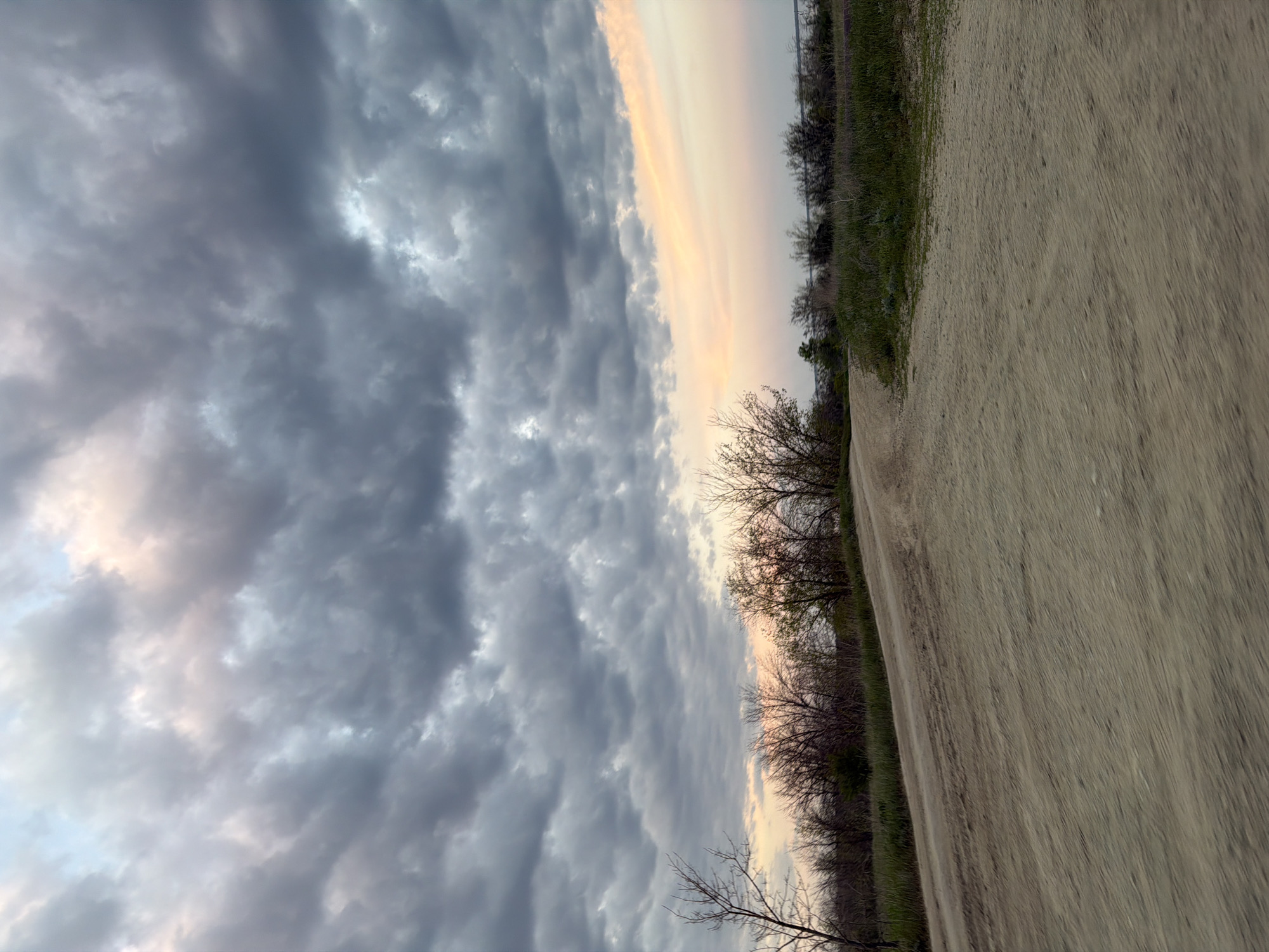 The image depicts a dirt path alongside a treeline under a cloudy sky at sunset. The path takes up the right portion of the frame, running diagonally from the lower right corner upwards, suggesting a downward slope. The treeline is on the left edge of the path, dividing the path from the sky. The sky occupies the left side of the frame and shows a sunset with dark, textured clouds dominating the view. A strip of orange light is visible between the clouds and the horizon near the treeline, indicating the setting sun. The location is Nizhnyaya Lipovka, Russia. There are no people visible.