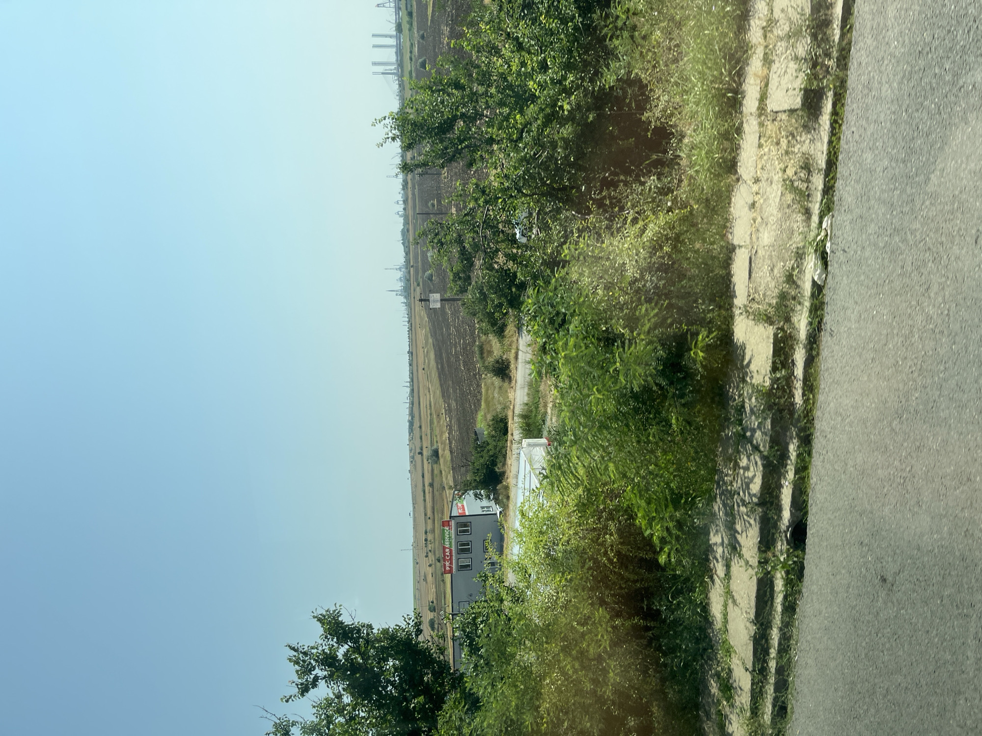 The image depicts an outdoor scene with a clear sky. In the foreground, a paved road is visible, bordered by a concrete sidewalk overgrown with grass and weeds. Beyond the sidewalk, a dense collection of green bushes and trees partially obscures the view.
Further back, a modern, rectangular building with a light-colored facade and several windows stands. A red and green sign above the building reads "PTC CAR SERVICE." Adjacent to this building, a white trailer or container is situated.
The midground of the image shows a cultivated field, with some areas appearing to be plowed or recently harvested, and other areas covered in dry vegetation. Scattered trees and utility poles are also present in this area.
In the distance, a sprawling industrial landscape is visible under a hazy sky. This includes numerous tall structures, cranes, and other industrial equipment, suggesting a port or factory complex. The overall impression is a landscape that transitions from a roadside with vegetation to agricultural land and finally to a distant industrial zone. The location is identified as Lazu, Romania, based on the provided context. There are no people visible in the image.