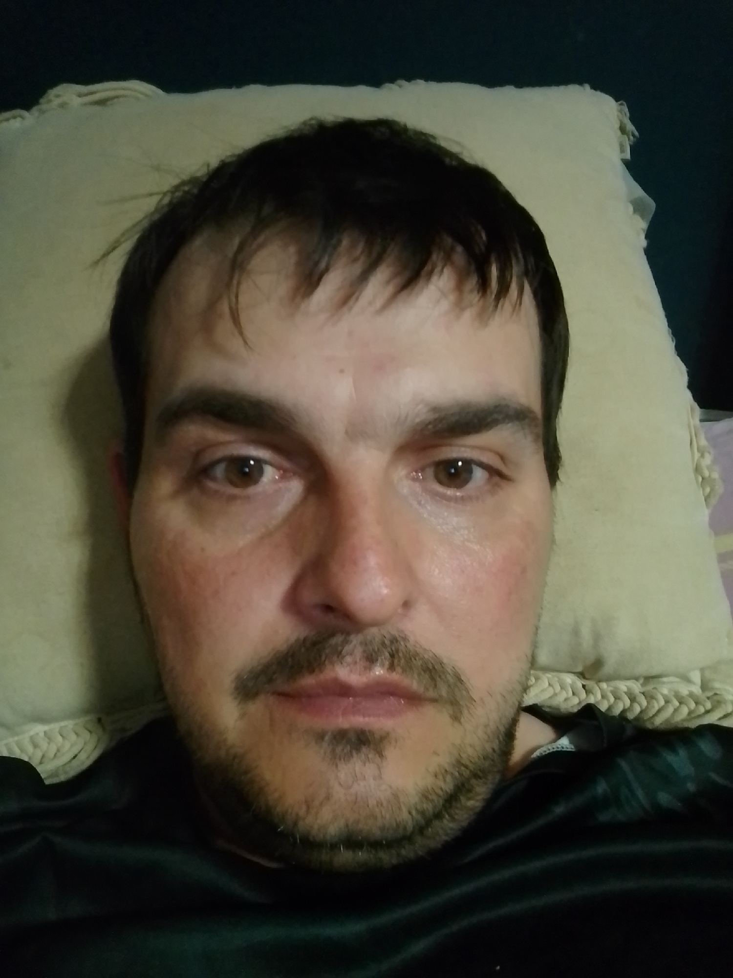 The image is a selfie of a Caucasian male lying down, with his head resting on a light-colored pillow. The background is dark. The man has dark hair, light brown eyes, a mustache, and a goatee. He is wearing a black shirt. The photograph appears to be taken indoors. There is no specific information in the image to indicate a location in Wedel, Germany.