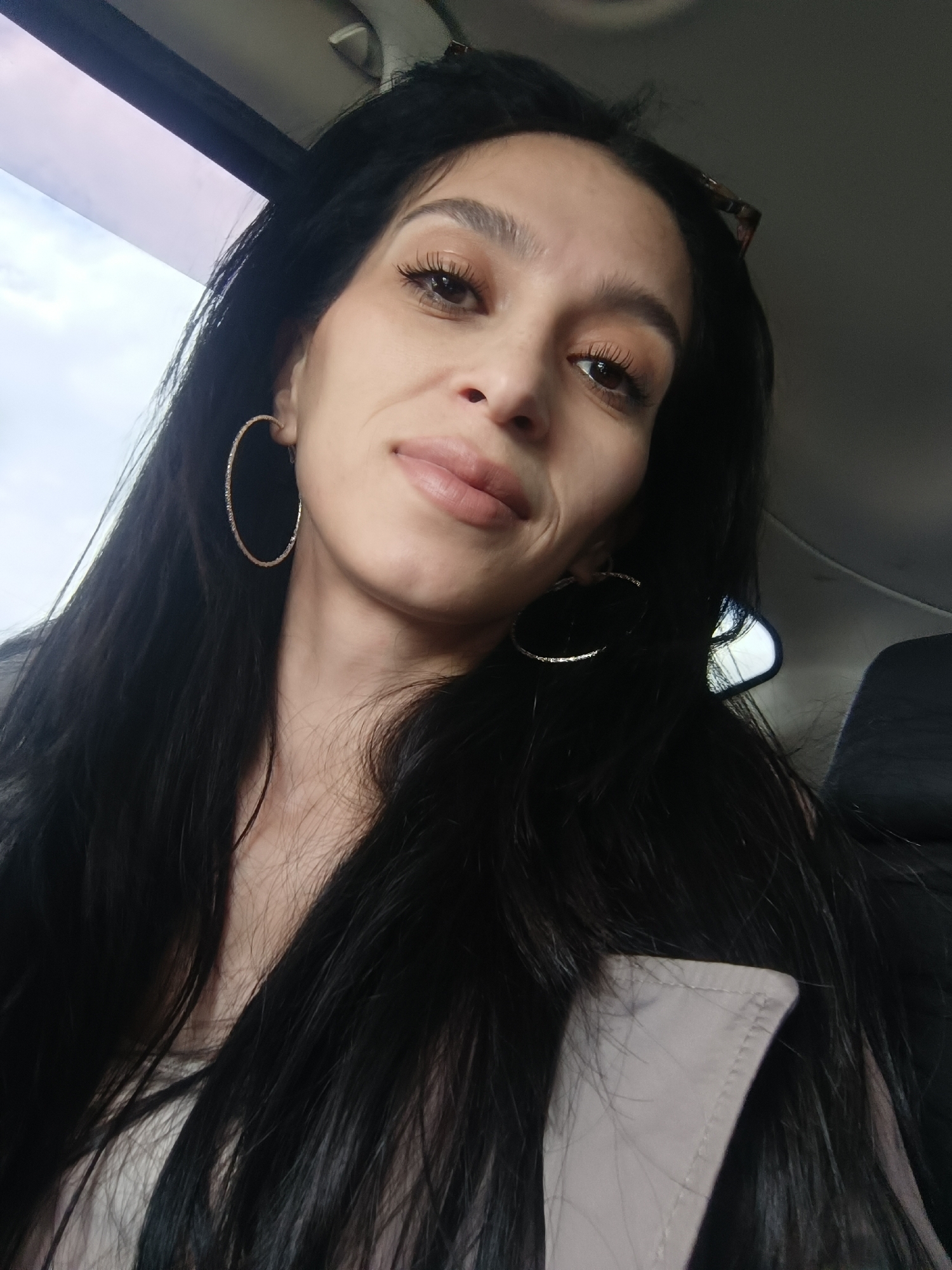 A woman is seated inside a vehicle, likely a car, based on the interior view. She has long, dark hair and is wearing large hoop earrings. Her makeup includes mascara and lip color. A piece of what appears to be a light-colored coat or jacket is visible in the lower right corner. The background shows an overcast sky visible through the car window. The photograph's location in the city of Укромновское сельское поселение, Ukraine, cannot be determined from visual information.