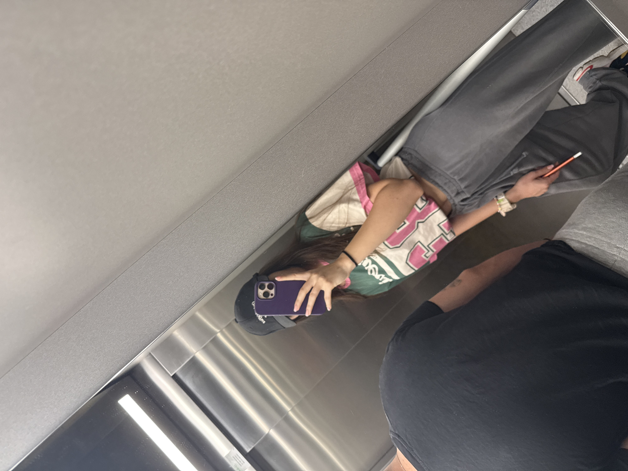 The image captures a tilted mirror selfie taken by a person in an indoor setting in Plovdiv, Bulgaria. The primary subject, reflected in a large, vertical metallic or mirrored surface, is a young person wearing a black baseball cap. They have long dark hair and are dressed in a casual outfit consisting of a colorful top with white, pink, and green patterns, and grey sweatpants. The subject is holding a purple smartphone up to take the photograph, with their hand and part of their arm visible. Their footwear, partially seen at the top right, appears to be sneakers with red and white details.
Partially visible on the right side of the reflection is another individual, seen from the waist down and side profile. This person is wearing a black t-shirt and grey shorts or pants, and is holding an orange pencil in their right hand. The reflective surface is framed by grey wall panels, and the reflections indicate overhead lighting and a sleek, modern interior, possibly an elevator or a hallway with stainless steel elements. No discernible text is visible on clothing or surroundings.