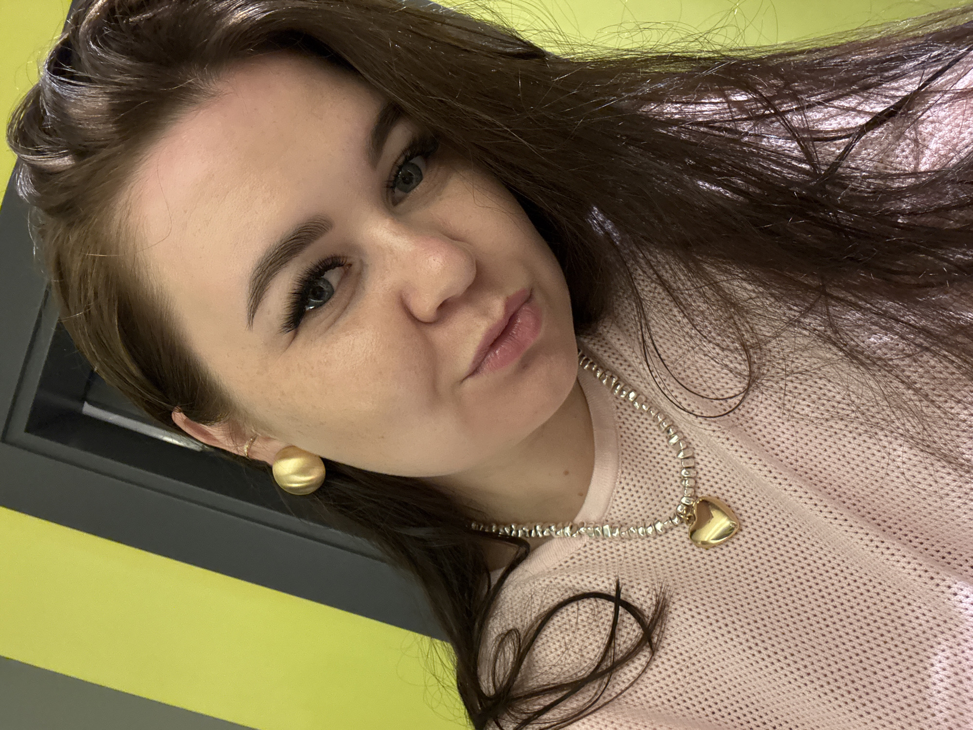 A selfie of a young woman with long, dark brown hair, fair skin, and blue eyes is shown. She is wearing a light pink, knitted sweater, a silver necklace with a gold heart pendant, and gold earrings. The background consists of a yellow and gray striped wall. The scene is indoors, with no visible indication of the city of Alanya, Turkey.