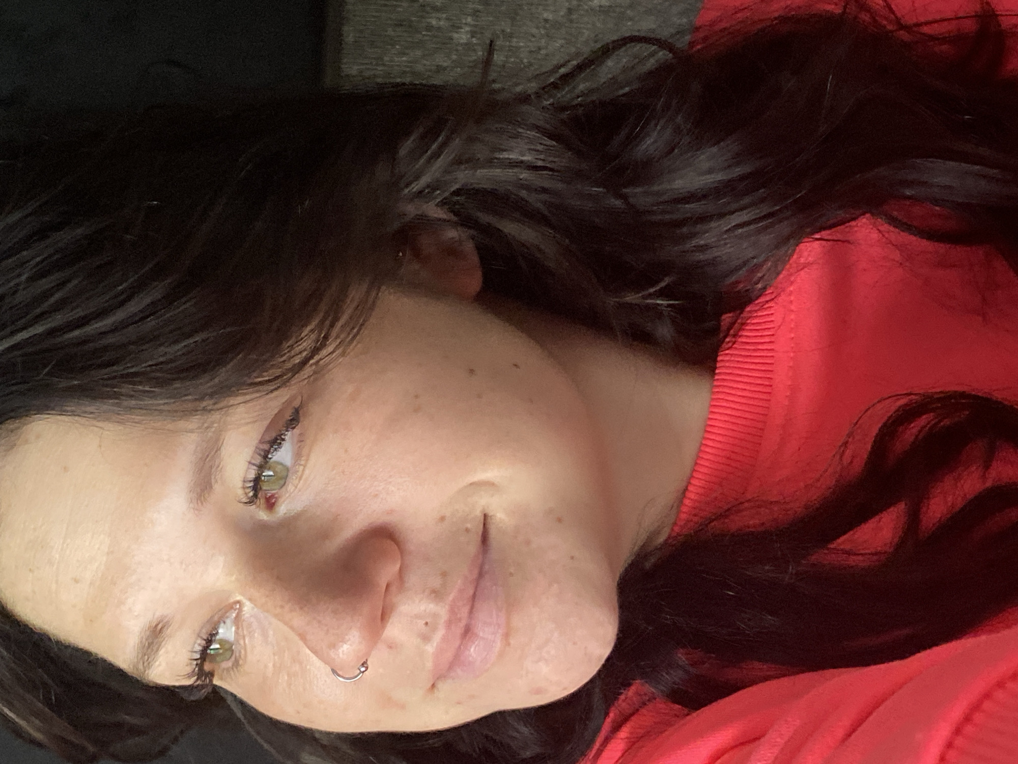 A close-up, profile view captures the right side of a young woman's face and shoulder. She has fair skin with scattered freckles and appears to be looking off to the left. Her eyes are a light green, and her lashes are dark and noticeable. She has a small silver hoop piercing in her right nostril. Her lips are a pale pink, slightly parted. Her dark brown hair, with natural waves, is visible around her face and shoulder. She is wearing a bright red, long-sleeved top with a ribbed neckline. The background is dark and indistinct, with a vertical textured element on the right side, possibly a wooden surface or wall. There is no clear indication of a specific location within the city of Chaikovsky, Russia.