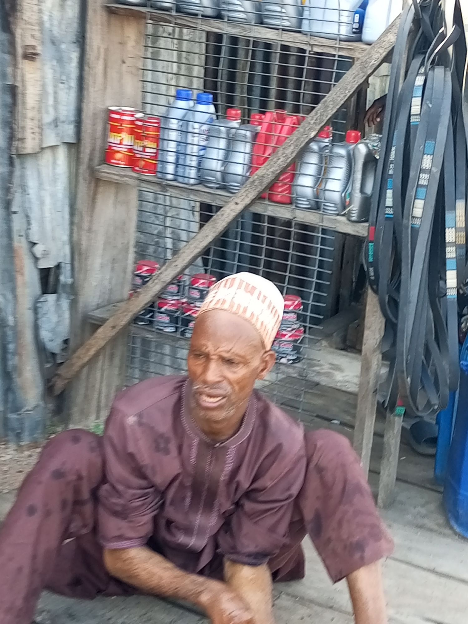 In Gombe, Nigeria, a man wearing a maroon robe and a patterned cap sits on the ground in front of a stall. The stall displays various items including red and silver cans of what appear to be automotive fluids and several black drive belts hanging vertically. The stall is constructed from metal grating and wooden planks, with a corrugated metal sheet forming the background. The overall environment suggests an outdoor market or repair shop setting. The man's expression is one of concern or discomfort as he looks towards the camera.