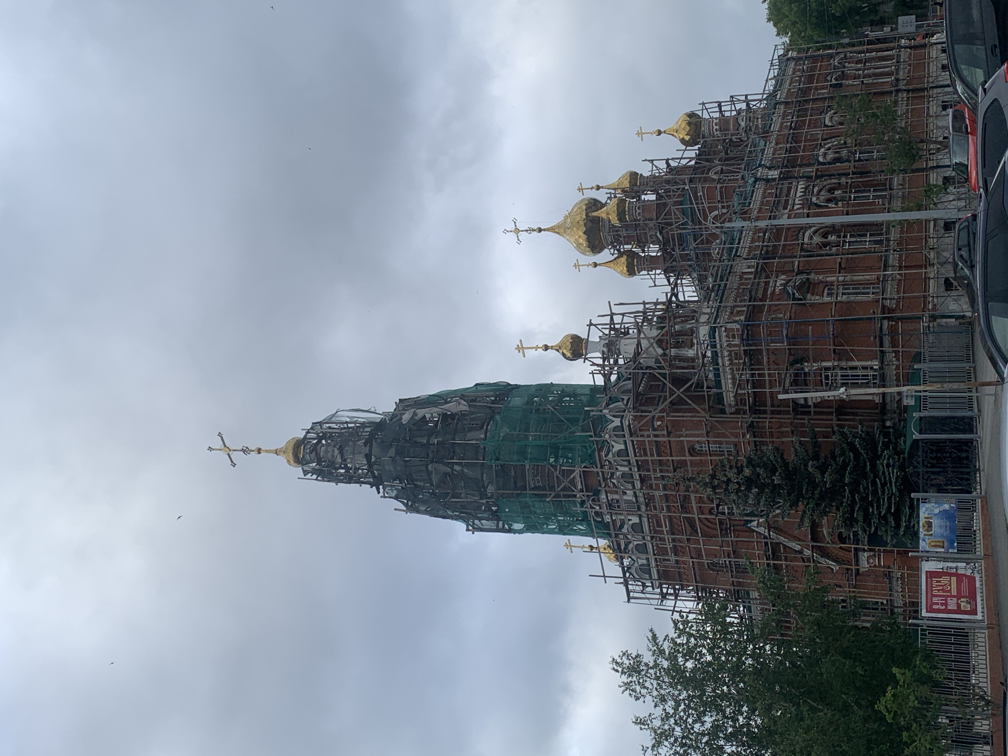 The image shows a large, red brick church with multiple golden domes and a prominent tall spire. The entire structure is covered in extensive scaffolding and green netting, indicating ongoing renovation or construction work. Several golden crosses are visible atop the domes and spire. In the foreground, there is a metal fence with some posters attached to it, and behind the fence, there are trees and a dark green fir tree. Several cars are parked along the street in front of the church. The sky is overcast and grey.
The church in the image is the Transfiguration Cathedral, located in the city of Perm, Russia.