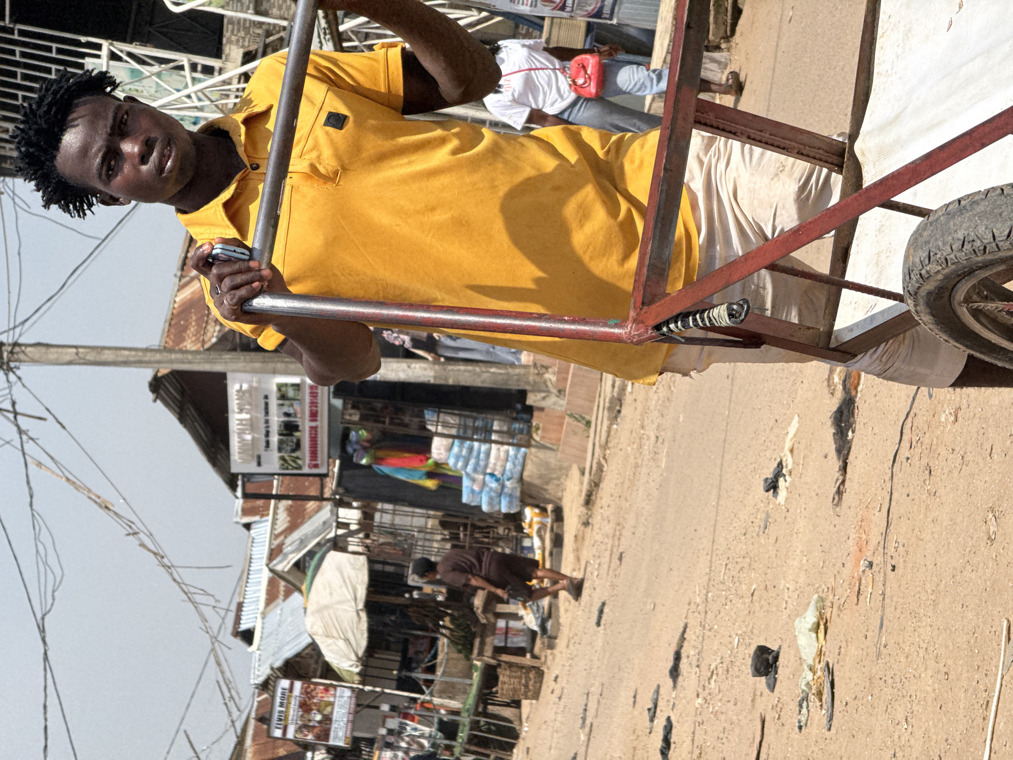 The image captures a street scene in Abuja, Nigeria. In the foreground, a young man with dark, dreadlocked hair is pushing a red metal cart. He is wearing a bright yellow polo shirt and light-colored shorts. His left hand is on the cart handle, and he holds a small blue device in his right hand. Behind him, the street is unpaved and dusty, with scattered debris. In the middle ground, several market stalls are visible, with corrugated metal roofs and signs. One sign reads "ELVIS MORE". People are seen walking in the background, including a woman wearing a white t-shirt and jeans, carrying a small red purse. The environment appears to be a bustling marketplace on a sunny day, with overhead wires and electrical poles suggesting an urban setting.