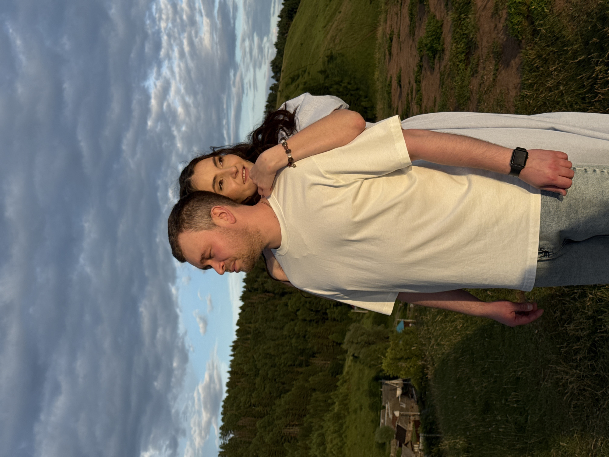 This media file shows a close-up of a couple in a rural outdoor setting during what appears to be golden hour. The image is oriented vertically, with the subjects in the foreground and a landscape in the background.
The main subjects are a man and a woman. The man, seen from the side and slightly from behind, has short brown hair and is wearing a white t-shirt and jeans. He has a watch on his left wrist. The woman is embracing him from behind, resting her head on his shoulder. She has long dark hair and is looking towards the camera with a gentle smile. She is wearing a light-colored garment. The focus is on the couple, with the background slightly blurred.
The setting appears to be a grassy hillside with a line of trees in the distance. There are some small buildings visible in the lower right portion of the image, suggesting a rural or village environment. The sky is filled with dramatic, cloudy formations, indicative of a setting sun.
The time of day is likely late afternoon or early evening, given the warm, diffused light and the cloudy sky. The overall mood conveyed is one of intimacy and affection between the couple. The location context provided is Olkhovka, Russia. There is no visible text in the image.