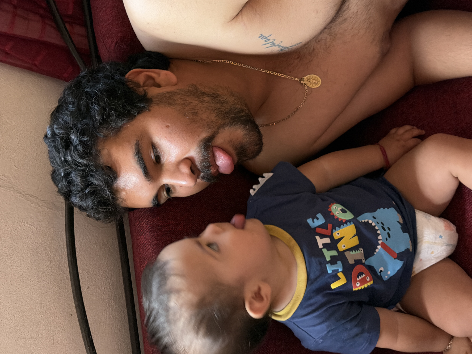 A man and a baby are lying face-to-face on a dark red, possibly burgundy, surface that might be a bed or couch. The man is topless, with a gold chain featuring a medallion around his neck and a tattoo visible on his upper chest. He has dark hair and a beard and is sticking his tongue out. The baby is wearing a navy blue shirt with a yellow collar and a dinosaur-themed design that includes the words "LITTLE DINOSAUR." The baby is also sticking its tongue out, mimicking the man. The baby is wearing a diaper. A black, vertical element, likely a bed frame, is seen on the left side of the frame. The scene is indoors. There is no evidence in the photograph to support the image being taken in Reforma, Mexico.