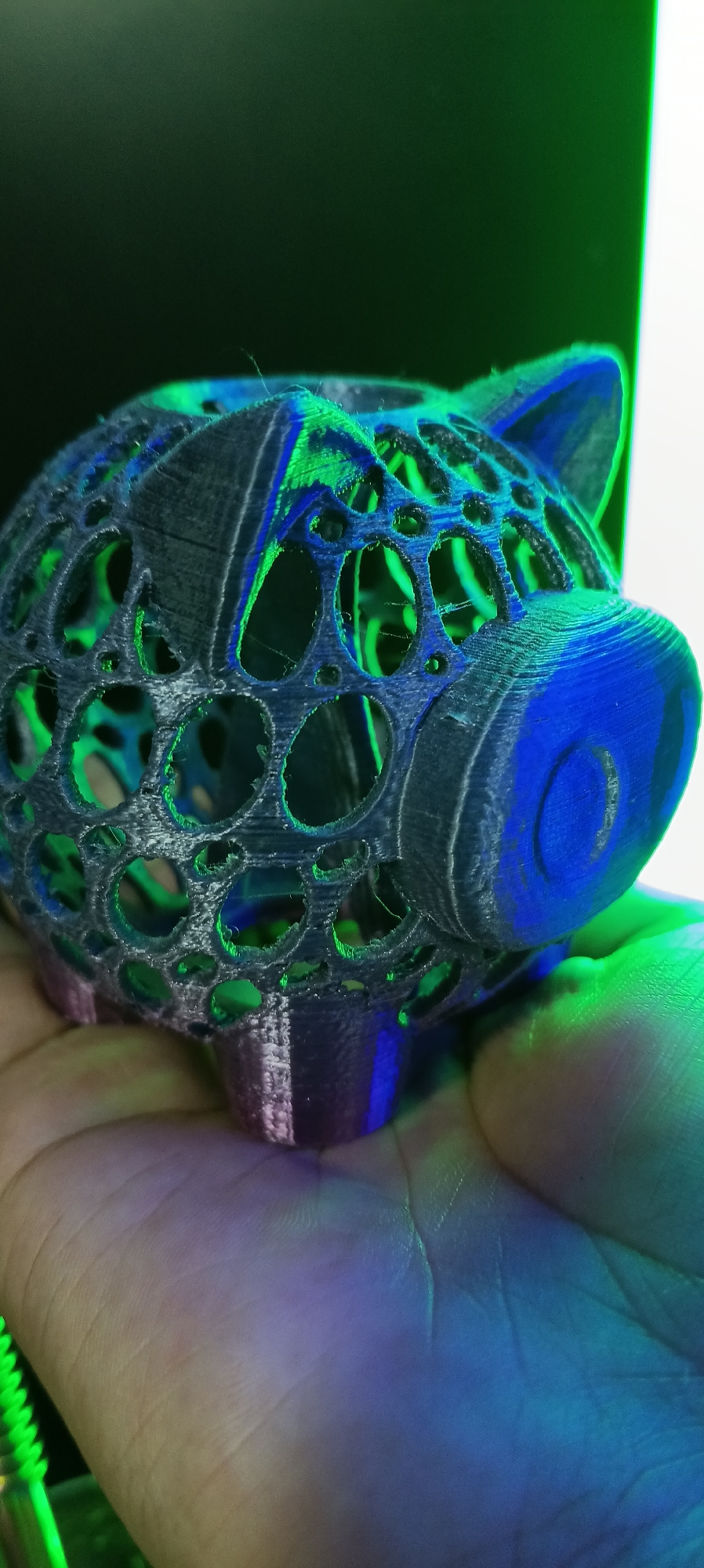A close-up shot captures a dark, 3D-printed piggy bank held in a person's palm, illuminated by vibrant green and blue lights. The piggy bank is intricately designed with numerous circular openings, revealing its hollow interior. The lighting casts dramatic shadows and highlights, emphasizing the texture and layered structure of the print. The background is a blend of dark and bright areas, suggesting an artificial lighting environment. The image does not contain information about the city of Wels, Austria.