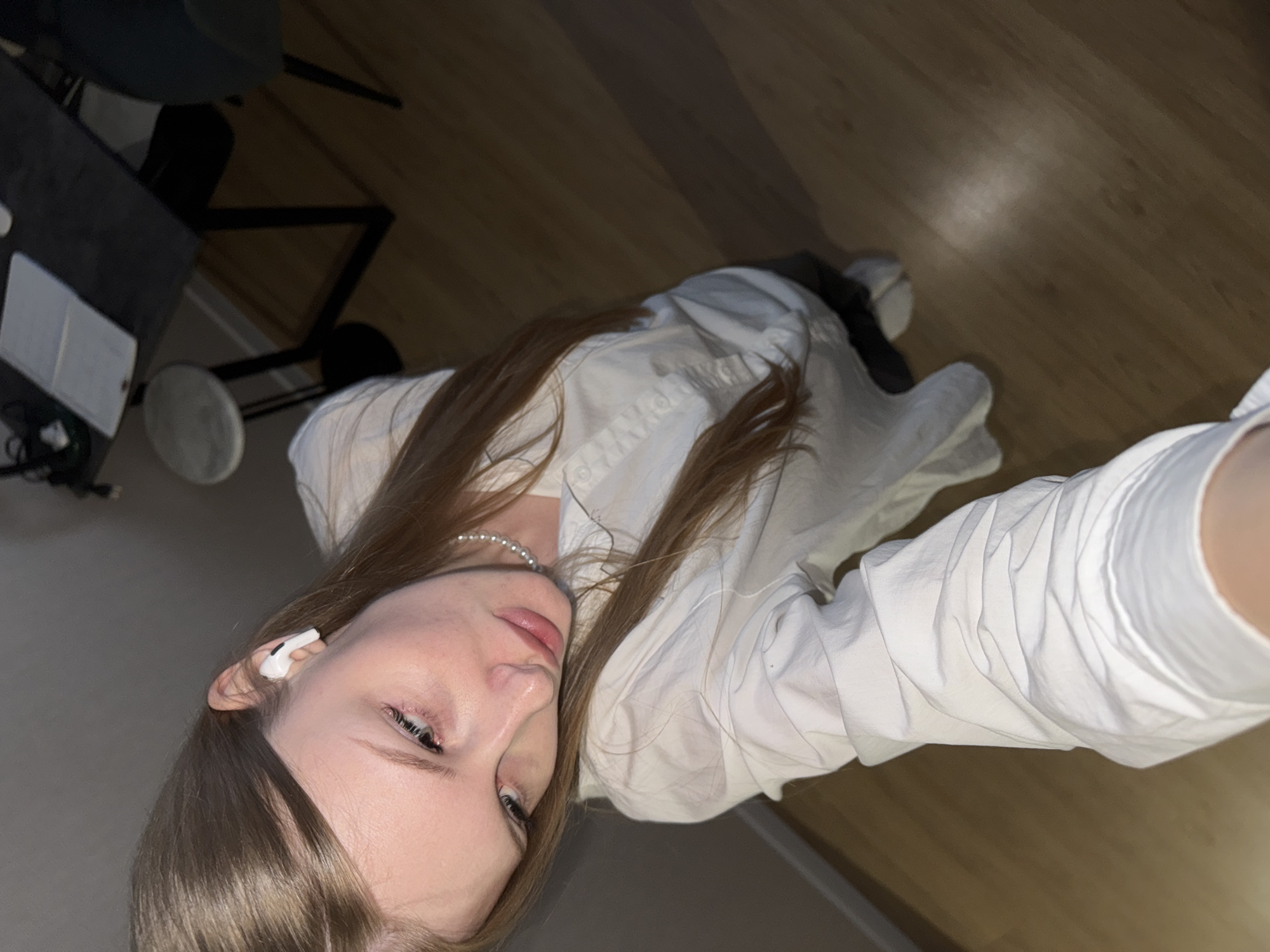This image, captured in Perm, Russia, features a young woman taking a selfie in an indoor setting. She has light brown hair and is wearing a white collared shirt and a pearl necklace. A white wireless earbud is visible in her right ear, suggesting she is listening to audio. Her gaze is directed downwards and to the left, with a neutral, somewhat introspective expression.
The background reveals a warm, light brown wooden laminate floor. To her left, part of a dark-colored table or desk is visible, holding a light-colored object, possibly a notebook. Nearby on the floor, there is a round, white object, potentially a speaker or part of a stand, along with some cables. Dark furniture legs are also partially visible in the background. The lighting in the room appears dim, suggesting either artificial illumination or that the photo was taken during the evening or night. There is no visible text within the image. The scene suggests a relaxed and personal environment.