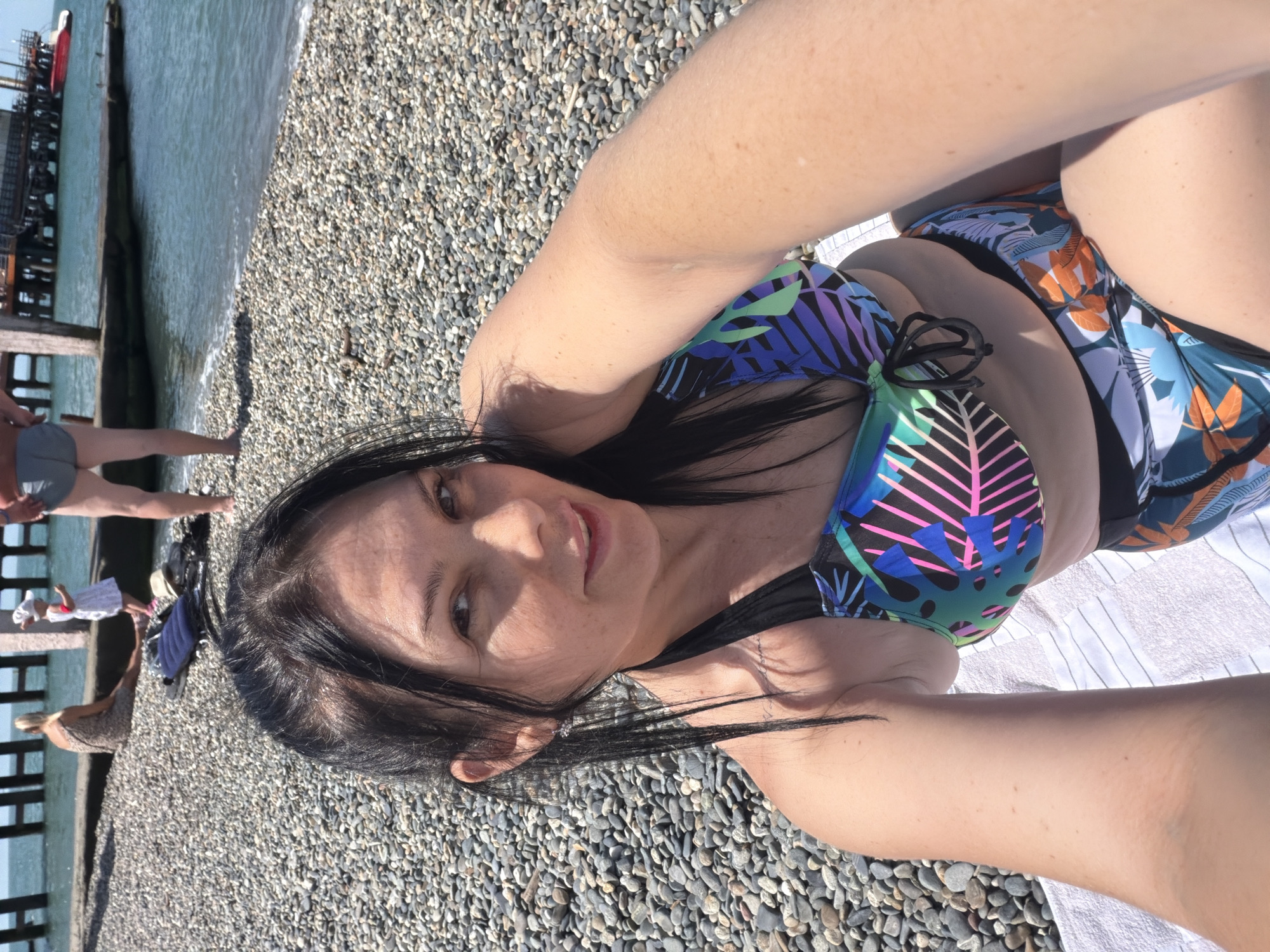 A woman in a colorful bikini is taking a selfie on a pebble beach. She is lying on a white towel. In the background, people are visible near a wooden pier over the Black Sea. The sea is calm and blue. The sky is clear and sunny. The location is identified as a beach in Sochi, Russia.