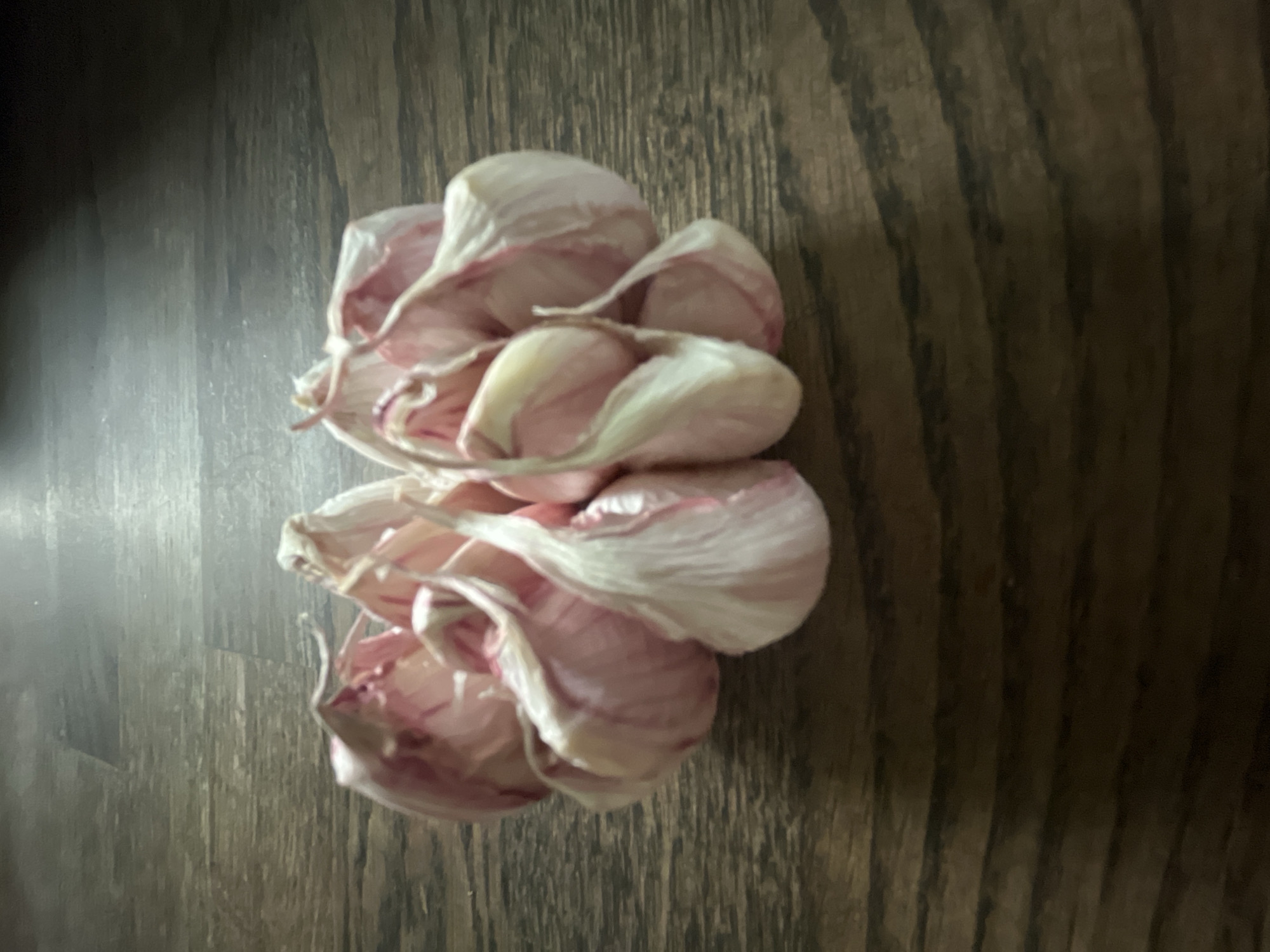 A head of unpeeled garlic lies on a dark, wooden surface with a visible grain pattern. The garlic has a mix of white and pinkish-purple coloration. The image shows a close-up, overhead shot. No specific city or location context is evident from the image.