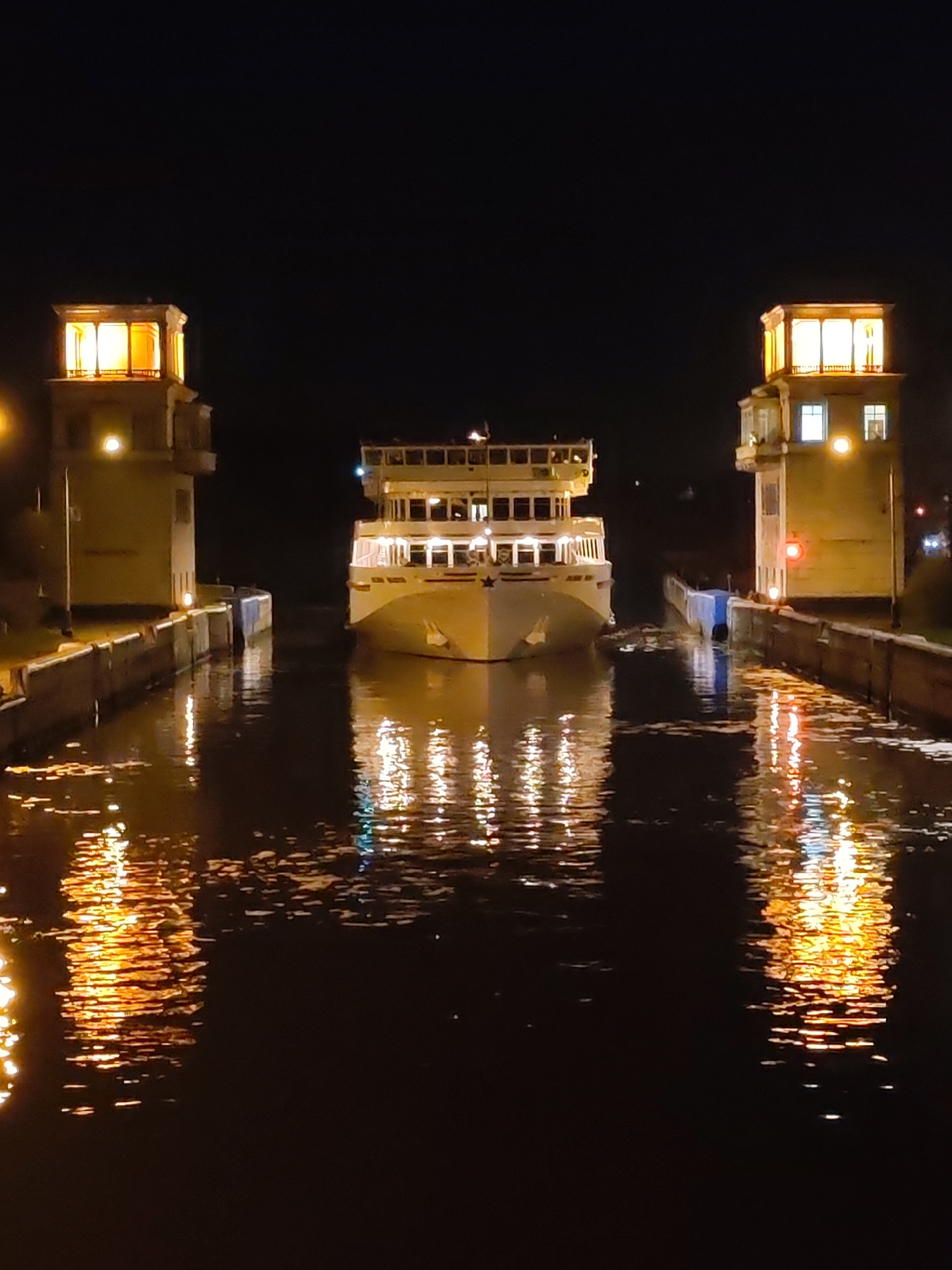A nighttime shot features a multi-story white passenger ship entering a lock on the Moscow Canal near Yakhroma, Russia. Flanking the ship are two illuminated towers on either side of the canal, with bright lights on top. Reflections from the towers and ship are visible on the water. The dark water of the canal fills the foreground and the background. The sky is dark and devoid of stars.