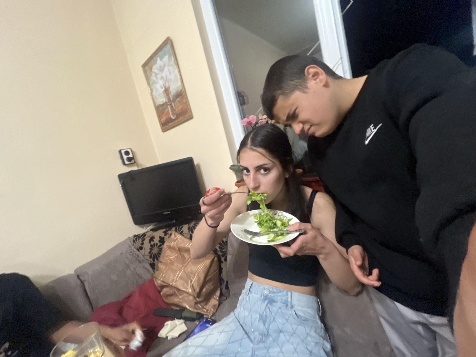 In what appears to be a living room, a young woman is eating a salad. She is wearing a black tank top and patterned light blue jeans. She is sitting on a grey couch with patterned throw pillows. To her right stands a young man in a black Nike sweatshirt. The room also contains a small television on a stand, a framed painting, and other unseen furniture. There is no discernible information in the image to conclude a location within Lozarevo, Bulgaria.