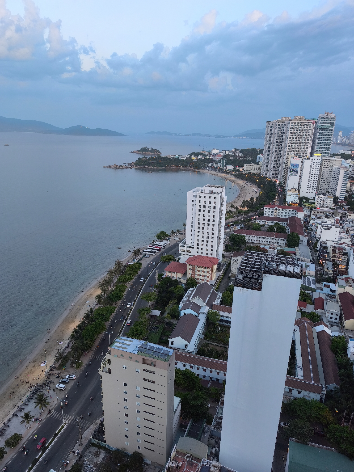 An aerial view captures the coastal city of Nha Trang, Vietnam at dusk. The scene is dominated by the vast, calm sea on the left, meeting a sandy beach dotted with numerous people and palm trees. A busy road runs parallel to the beach, lined with cars, motorcycles, and buses, indicating ongoing activity.
The cityscape features a mix of modern high-rise buildings, some quite tall and densely packed, interspersed with lower-rise structures and lush greenery. One prominent white, rectangular building stands out, its sheer face reflecting the sky. Another tall, light-colored building with many windows is visible closer to the beachfront.
In the distance, several islands are silhouetted against the horizon, with a few structures visible on the closest one. The sky is filled with soft, layered clouds in shades of blue and gray, typical of twilight. The overall impression is one of a vibrant coastal city at the end of the day. There is no discernible text in the image.