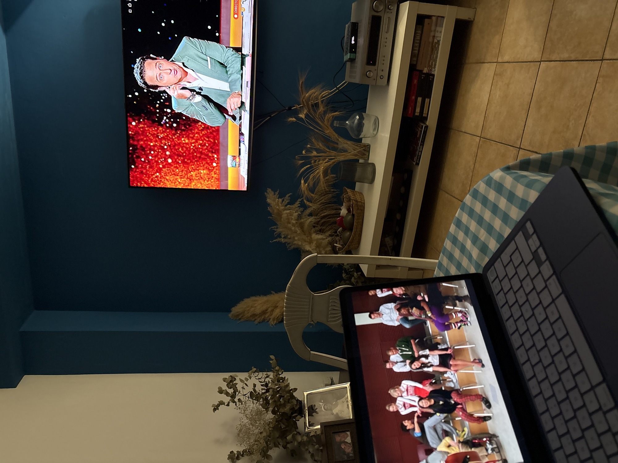The image captures an indoor setting, likely a living room, within Urbanización Encinares De Alcolea, Spain. The scene features a large, wall-mounted flat-screen television on a dark teal wall, displaying a man in a light green suit jacket, appearing animated against a background of sparkling orange and red lights. Below the TV, a white shelving unit holds an audio system, books, and various decorative items including dried plants and vases. To the right, a laptop rests on a surface covered with a blue and white checkered blanket or tablecloth, its screen showing a group of people seated in what appears to be a classroom or audition setup.
The primary activity depicted is simultaneous media consumption: someone is watching a program on the large television and engaging with content on the laptop. The man on the TV seems to be a presenter or performer in a live broadcast.
Notable details include the contrasting dark teal wall with a lighter upper section and ceiling, light brown tiled flooring, and ambient lighting that suggests evening or nighttime. The decorative elements, such as dried grasses, multiple vases, and framed pictures near the floor, contribute to a homey atmosphere.
Visible text includes "DIRECTO" (Spanish for "LIVE") on the large TV screen, a small "HD" icon, and potentially a channel logo like "LA 1". On the audio system, "gms." is visible. The spine of a book on the shelf clearly shows "FOLLETT".