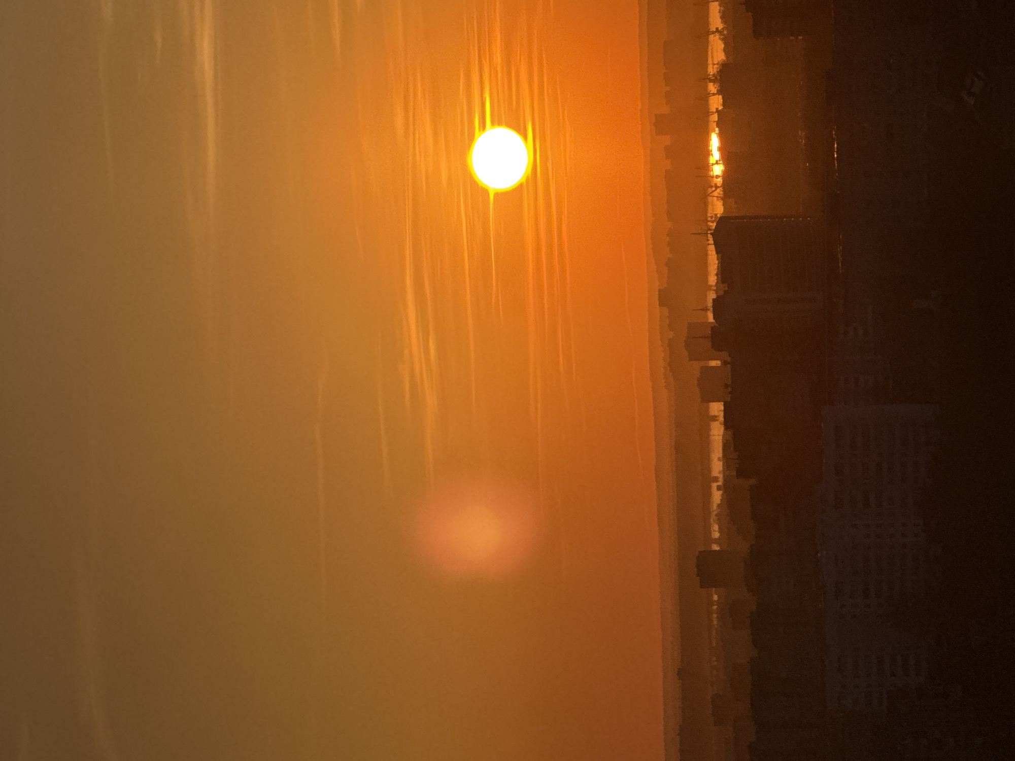 A photograph shows a hazy, golden sunrise or sunset over a city landscape. The bright sun is visible near the top of the image, with a blurred reflection of the sun appearing below it. To the right, the silhouetted outlines of buildings and structures indicate a city skyline. The sky is filled with a warm, orange glow. Based on the buildings, this is not Perm, Russia.