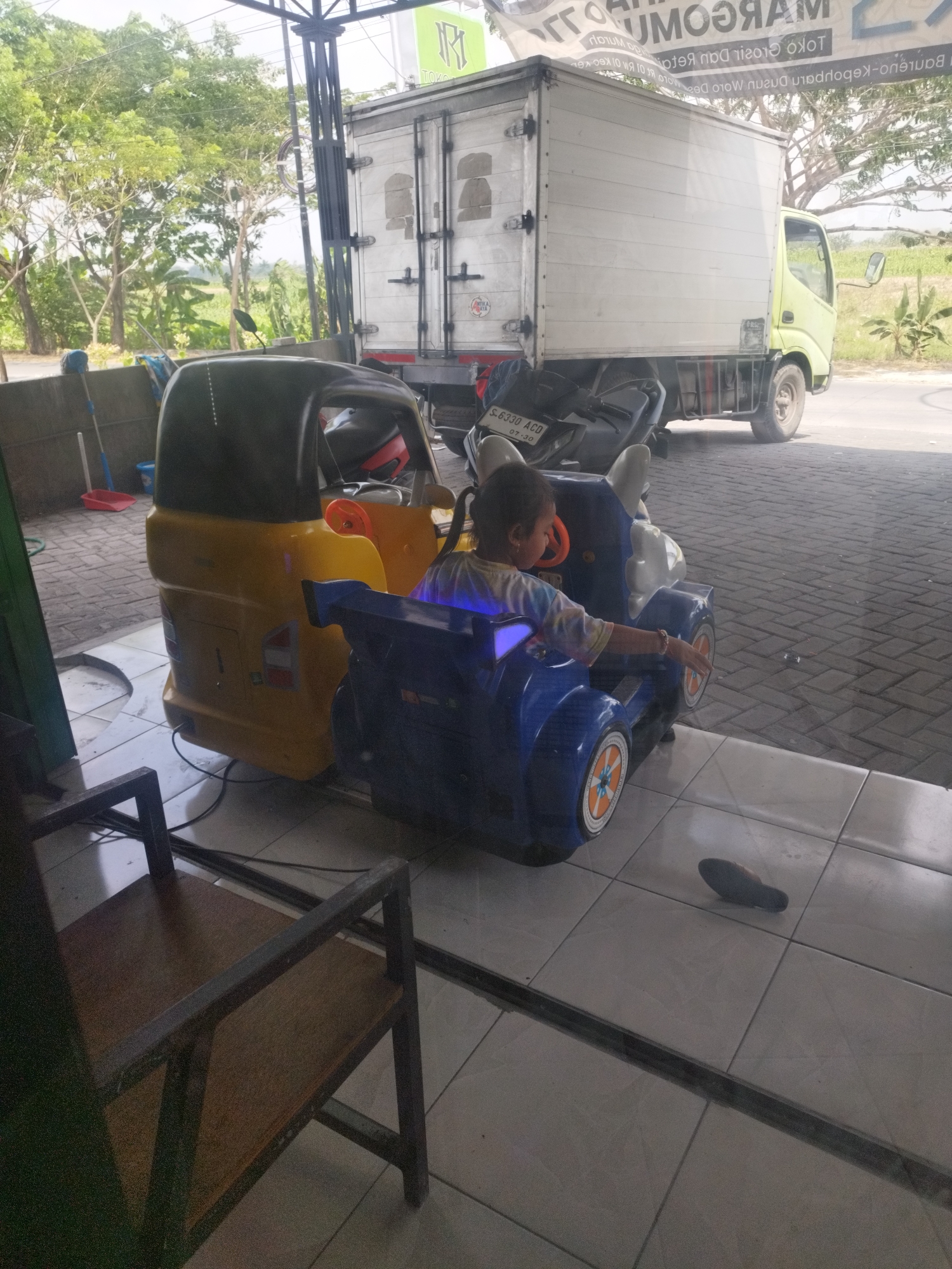 In Woro, Indonesia, a young child is seated in a blue toy car with a black seat, positioned behind a yellow ride-on vehicle. The child is wearing a tie-dye shirt. The toy cars are on a tiled floor under a covered structure. Behind the toys, a white truck with a light yellow cab is parked. To the left of the yellow toy vehicle, a red dustpan and a blue bucket are visible. Trees and foliage are present in the background, and a street is visible in the distance.