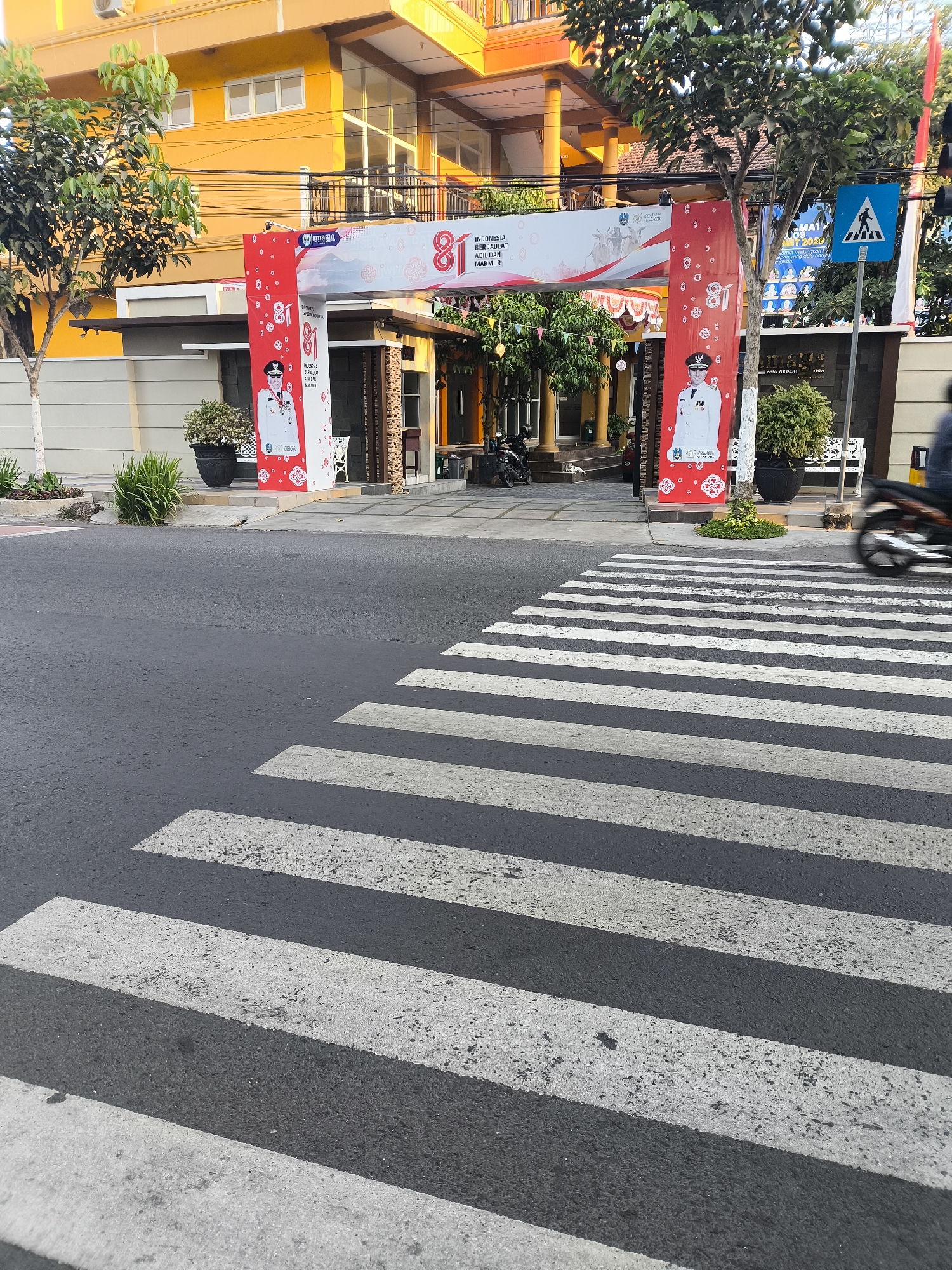 The image shows a pedestrian crossing on a street in front of a building with an orange facade. An arched banner is set up over the entrance to the building, decorated with red and white fabric and featuring the number "81" prominently. Two large red banners hang from the arch, each displaying a portrait of a man in a uniform and text in Indonesian. The building appears to be a school or public institution, with trees and potted plants in front. A motorcycle is visible in the background, and the street is paved with asphalt. A pedestrian crossing sign is mounted on a pole to the right of the banner.