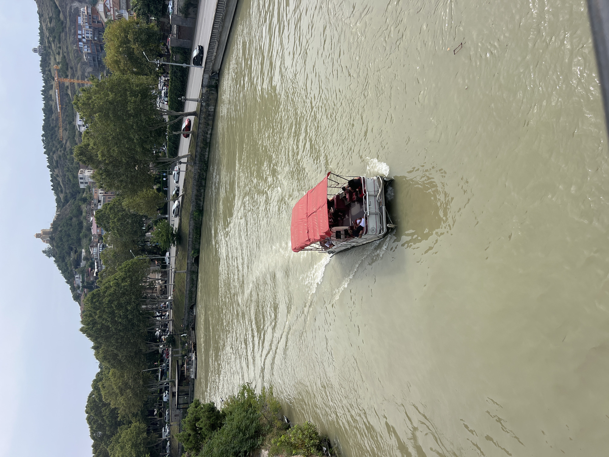 In Tbilisi, Georgia, a scenic view captures the Mtkvari River, with a boat carrying tourists sailing through its murky waters. The boat, adorned with a red canopy, is seen from an elevated perspective, likely from a bridge, as it navigates the current. On the riverbank, lush green trees line the path, with a road bustling with traffic and cars in the background. In the distance, buildings and homes dot the hillside, leading up to a prominent church crowning the summit. The sky overhead is overcast, casting a soft light on the urban landscape.
