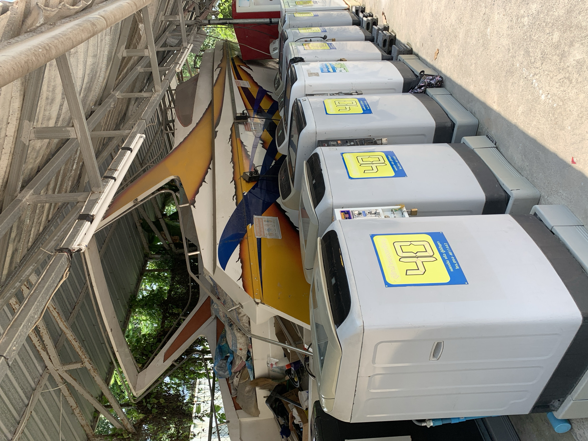 The image captures an outdoor or semi-sheltered service area, featuring a row of coin-operated washing machines alongside a large boat. The setting appears to be under a corrugated metal canopy supported by a metal frame structure.
In the foreground and extending into the midground, numerous light gray, top-loading washing machines are neatly lined up. Each machine features a yellow and blue sticker with Thai text. These stickers indicate "ตู้ซักผ้าหยอดเหรียญ" (Coin-operated washing machine), the number "40" (possibly a price or machine number), and contact information "แลกเงิน หรือ ตู้มีปัญหา โทร.094-4011442" (Exchange money or machine has problems, Tel. 094-4011442). One machine has a small, patterned bag resting on it.
To the left of the washing machines, partially obscured by the metal canopy structure, is a distinctive boat. It features a vibrant design with yellow, blue, black, and white sections, suggestive of a speed boat or a long-tail boat. It appears to be stored or undergoing some work, with various items and clutter visible around its base. Green foliage and trees are visible in the background, suggesting a natural environment surrounding the service area.
The scene is brightly lit by daylight, casting strong shadows on the concrete ground next to the washing machines, indicating a sunny day. The overall impression is that of a functional, practical service facility, possibly located near water given the presence of the boat, in บ้านบางรักษ์, Thailand.