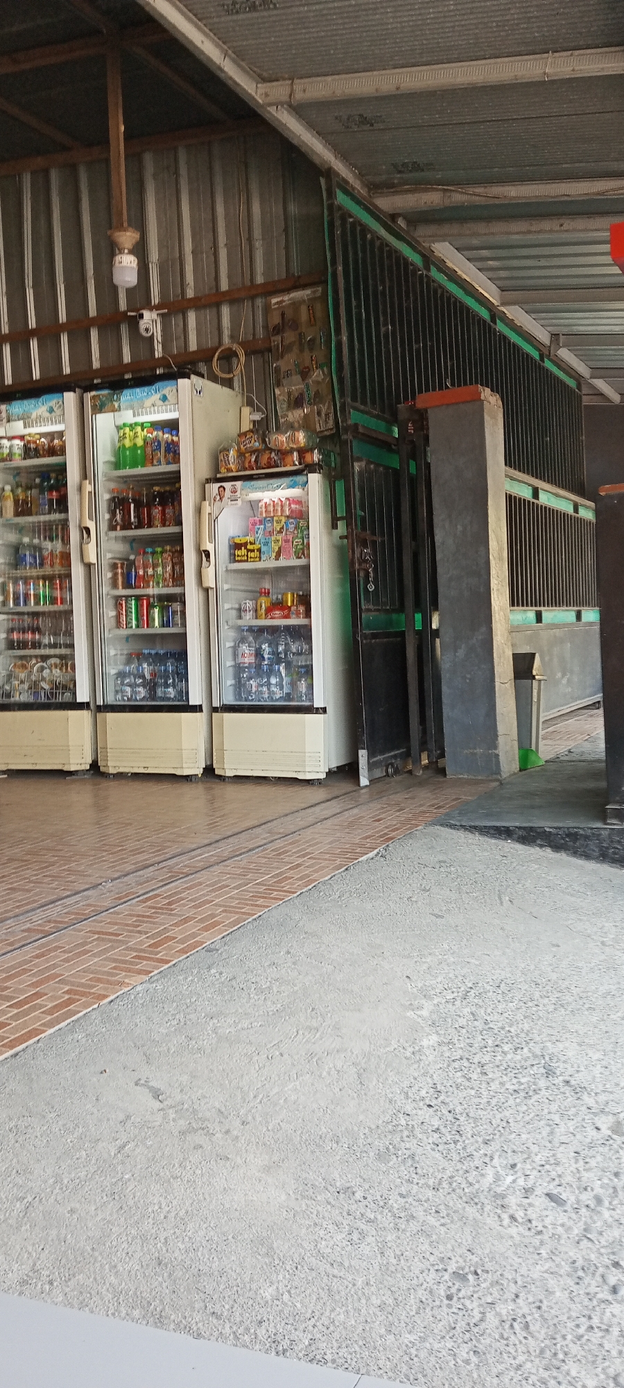 In the city of Pakkanna, Indonesia, a ground-level view captures the exterior of a small shop or convenience store. The focal point is a row of three glass-doored refrigerators filled with beverages, standing side-by-side. To the right of the refrigerators, a metal gate with green accents is partially open, revealing a concrete pillar and a trash can. The ground is a mix of tiled flooring in a brick pattern and rough concrete. Above, the corrugated metal roof of the establishment is visible, supported by wooden beams. A security camera is mounted on one of the beams. The scene appears to be in daylight, with the sunlight creating shadows and highlighting the textures of the environment.