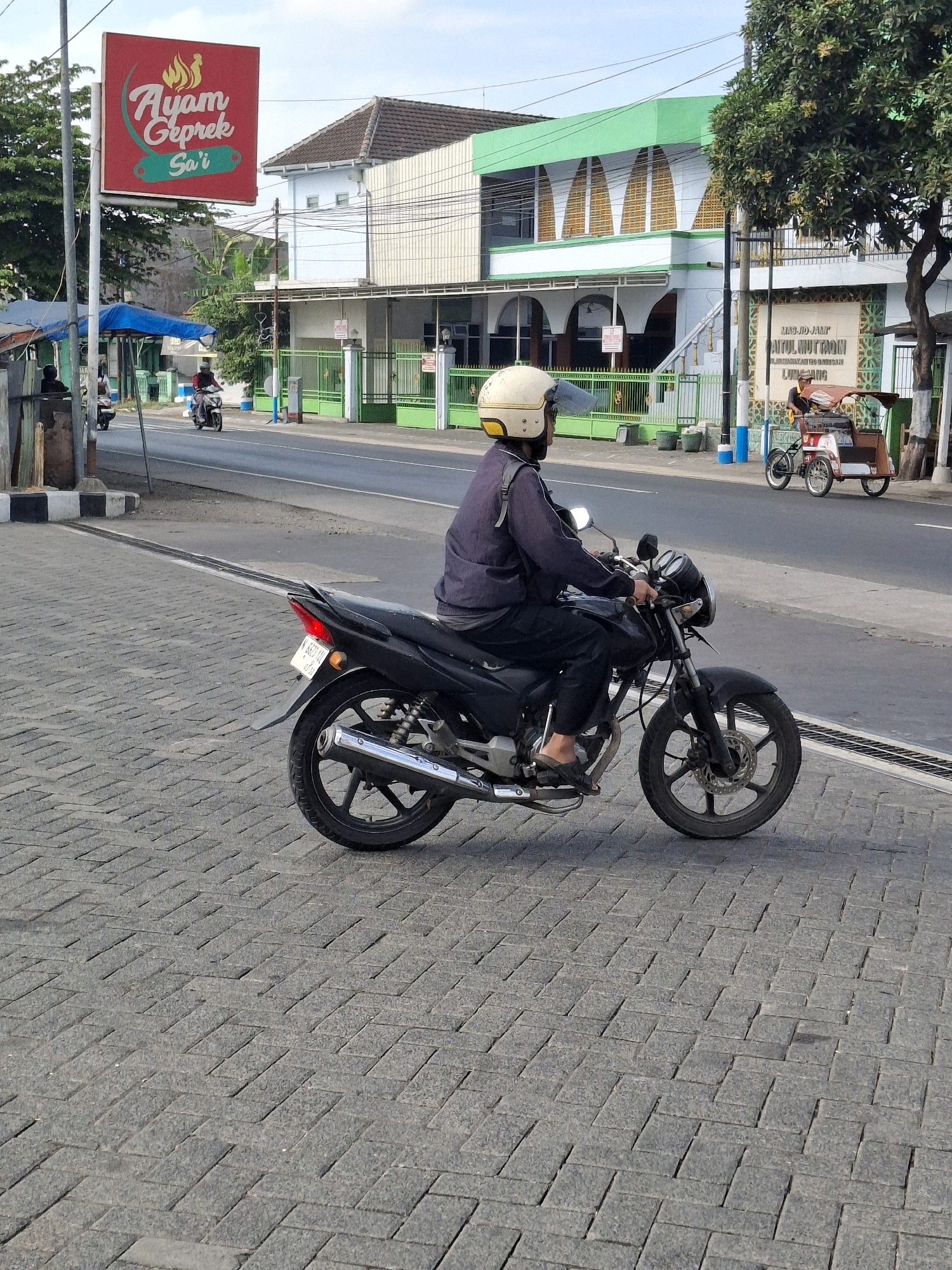 In Lumajang, Indonesia, a person wearing a yellow helmet rides a black motorcycle on a brick-paved sidewalk adjacent to a street. The rider is dressed in dark clothing. In the background, across the street, are buildings and a sign that reads "Ayam Geprek Sa'i." Further down the street, a motorized tricycle is parked near a building with green accents, possibly a mosque, with signage indicating "MASJID JAMI' SYAITUL MUTTAQIN." The sky is partly cloudy.