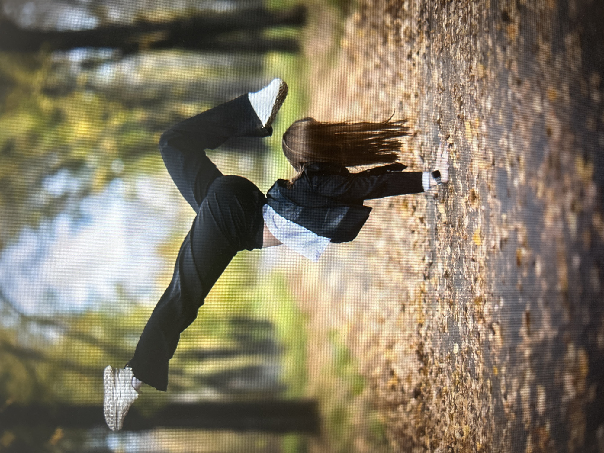 A young woman with long brown hair is performing a handstand on a path covered with fallen autumn leaves. She is wearing black pants, white sneakers, a white shirt, and a black jacket. Her body is positioned vertically, supported by her hands, with her legs split in the air. The background is a blurred forest with trees and dappled sunlight. The location of this scene is not specified in the image or the provided context. Therefore, it is impossible to determine if it is in Kiselyovsk, Russia, or any other city.