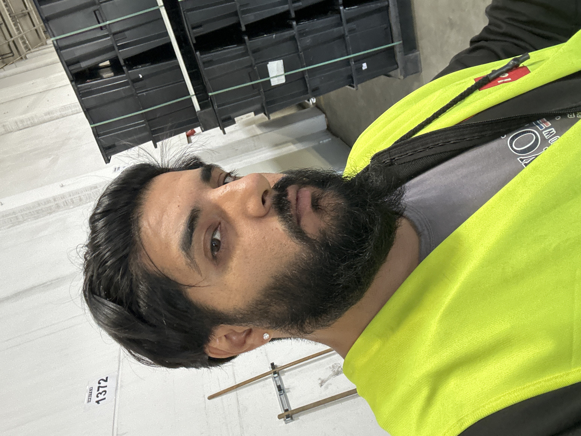 A man with dark hair and a beard, wearing a grey t-shirt and a bright yellow safety vest, stands in a warehouse. He has a diamond stud earring in his left ear. Behind him, stacks of black plastic bins are visible. To his left, a white wall has the number "1372" printed on it, and a wooden broom is leaning against the wall. The floor appears to be concrete. The scene is well-lit.