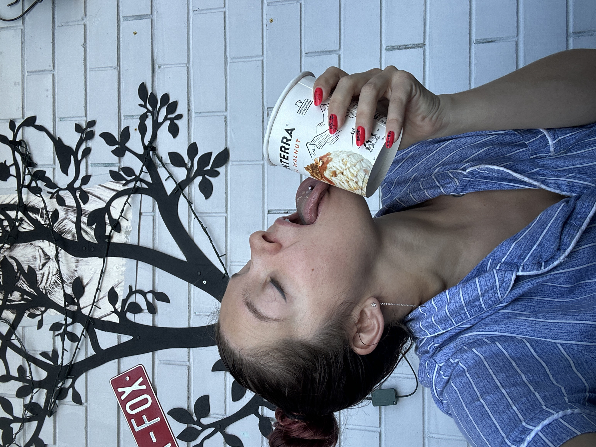 The media file shows a young woman with her eyes closed, licking ice cream from a paper cup. She is wearing blue and white striped pajamas. The background is a white brick wall with a black metal tree decoration. A small red sign with white text that reads "FOX" is visible. The woman has red nail polish and a silver necklace. A small silver star earring is visible in her ear. The lighting suggests it is daytime. The overall mood is playful and suggestive.