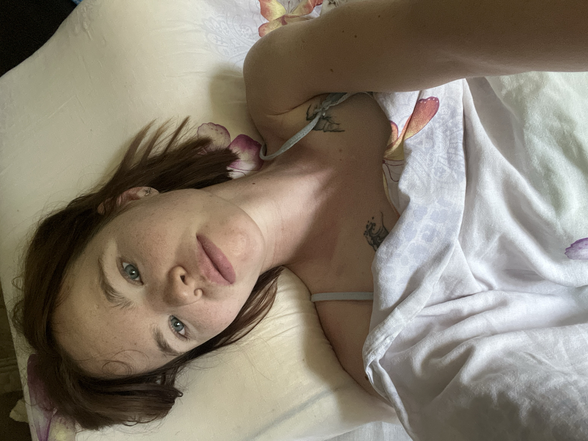 A young woman with medium-length brown hair lies on a pillow, looking directly at the camera. She has light skin with freckles and blue-green eyes. Her lips are slightly parted. She is wearing a light-colored spaghetti strap top and is covered by a white comforter with a floral pattern. A tattoo of a wave is visible on her upper chest, and another tattoo, possibly of a spider, is visible on her shoulder. The background is a bed with a floral patterned pillow and comforter. The lighting suggests an indoor environment. There are no obvious indicators of the specific location within Chaikovsky, Russia.