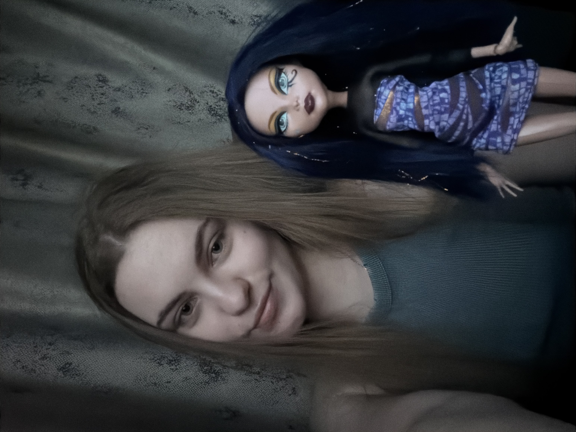 A young woman with long, blonde hair, wearing a teal ribbed tank top, is posing next to a Monster High doll with long, dark blue hair and distinctive makeup. The doll is dressed in a purple and blue patterned strapless dress with black mesh sleeves and accents. The background appears to be a dark, textured curtain. The image was taken indoors, with the lighting suggesting a casual selfie. There is no discernible information indicating the location in Dnipro, Ukraine.