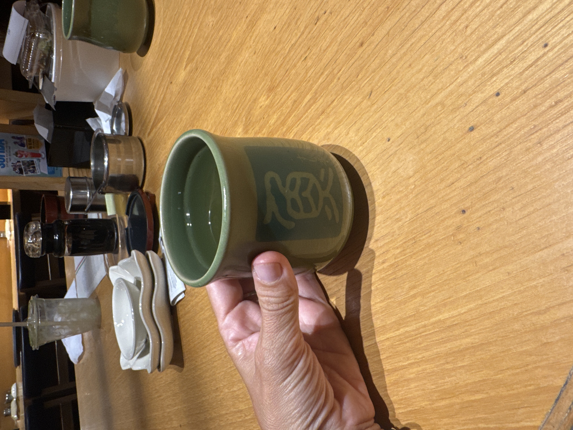 The image captures an indoor scene, likely a restaurant or cafe, with a wooden table serving as the primary surface. The dominant subject is a human hand, appearing to be an adult's, holding a green ceramic mug. The mug has a light green or tan patterned design on its side, which abstractly resembles a turtle or stylized motif.
Various other dining-related objects are scattered across the wooden table in the background. These include another green mug, a light-colored bowl or container, several shiny metallic ramekins or serving dishes, and a stack of white and cream-colored ceramic dishes, some with a distinct wavy or floral edge. A clear glass or plastic cup with a straw is also visible, along with a dark bottle that might be a condiment dispenser. Crumpled white paper or napkins are present on the table.
In the far background, partially obscured, is a blue and white poster or sign. The word "SOUDA" is clearly visible on this sign, with other smaller, unreadable text below it. The lighting suggests an indoor setting, possibly daytime, but could also be well-lit artificial illumination. The image is oriented sideways. The scene depicts a moment in a dining environment in Kuta Selatan, Indonesia.