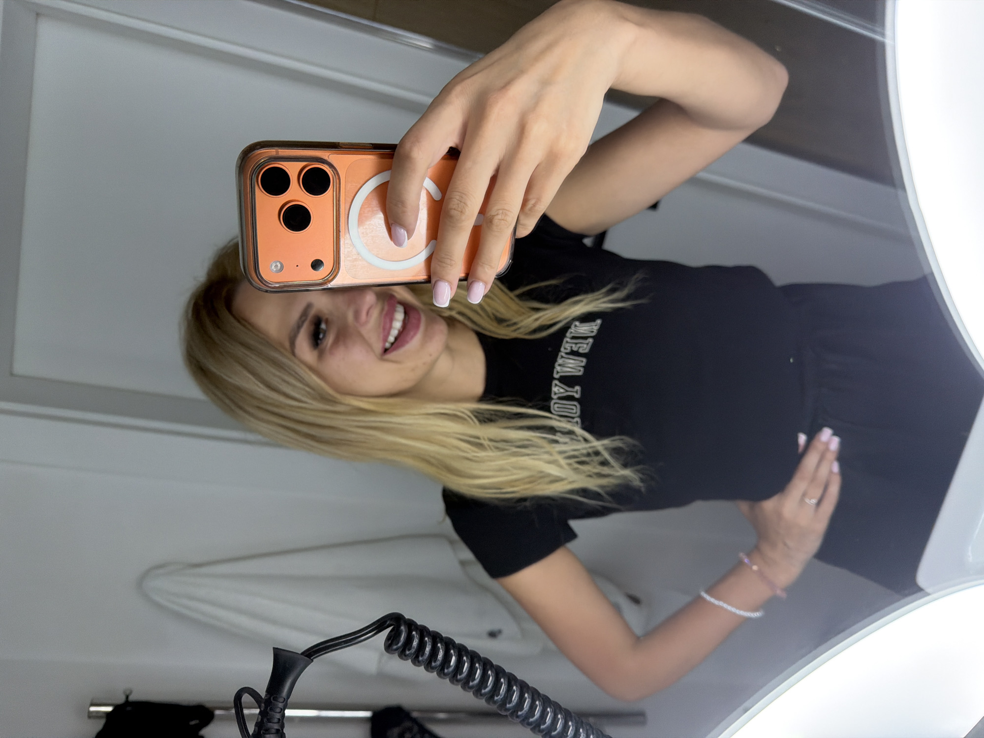 A blonde woman is taking a selfie in a bathroom. She is wearing a black t-shirt with white lettering that appears to say "NEW YORK". She is holding an orange iPhone in her right hand, which is reflecting in the mirror. She has her left hand on her hip. The bathroom appears to be modern with bright lighting, and a towel or robe is hanging on the wall in the background. The scene is indoors and appears to be a personal space. There is no information in the image to indicate the location in Polianytsia, Ukraine.
