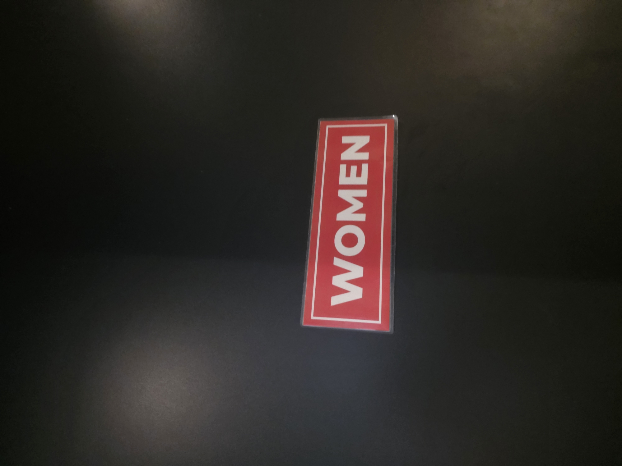 A rectangular sign with a red background and a white border displays the word "WOMEN" in large white capital letters. The sign is affixed to a dark, likely matte, surface, which could be a wall or door. The lighting in the image is dim, creating shadows and highlighting the sign. The specific location in North Kuta, Indonesia, is not discernible from the image itself, as there are no identifying landmarks or contextual clues. The scene is static, with no people or activity visible.