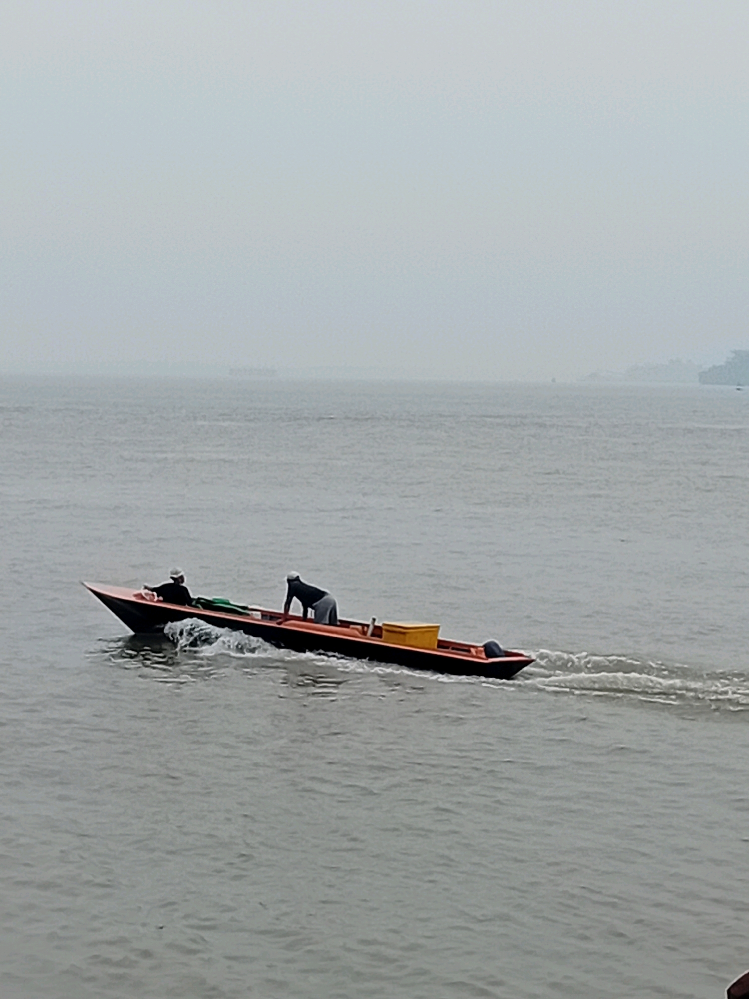 A long, narrow motorboat, painted black with an orange trim and containing a yellow container, moves through a body of water, leaving a wake. Two individuals are on board. One person, wearing a white cap and dark clothing, sits at the front of the boat. Another person, in dark clothing and light gray pants, stands at the back. The boat is traveling from left to right. The background is dominated by a hazy sky and a distant, indistinct shoreline or landmass. The overall atmosphere is overcast and muted.