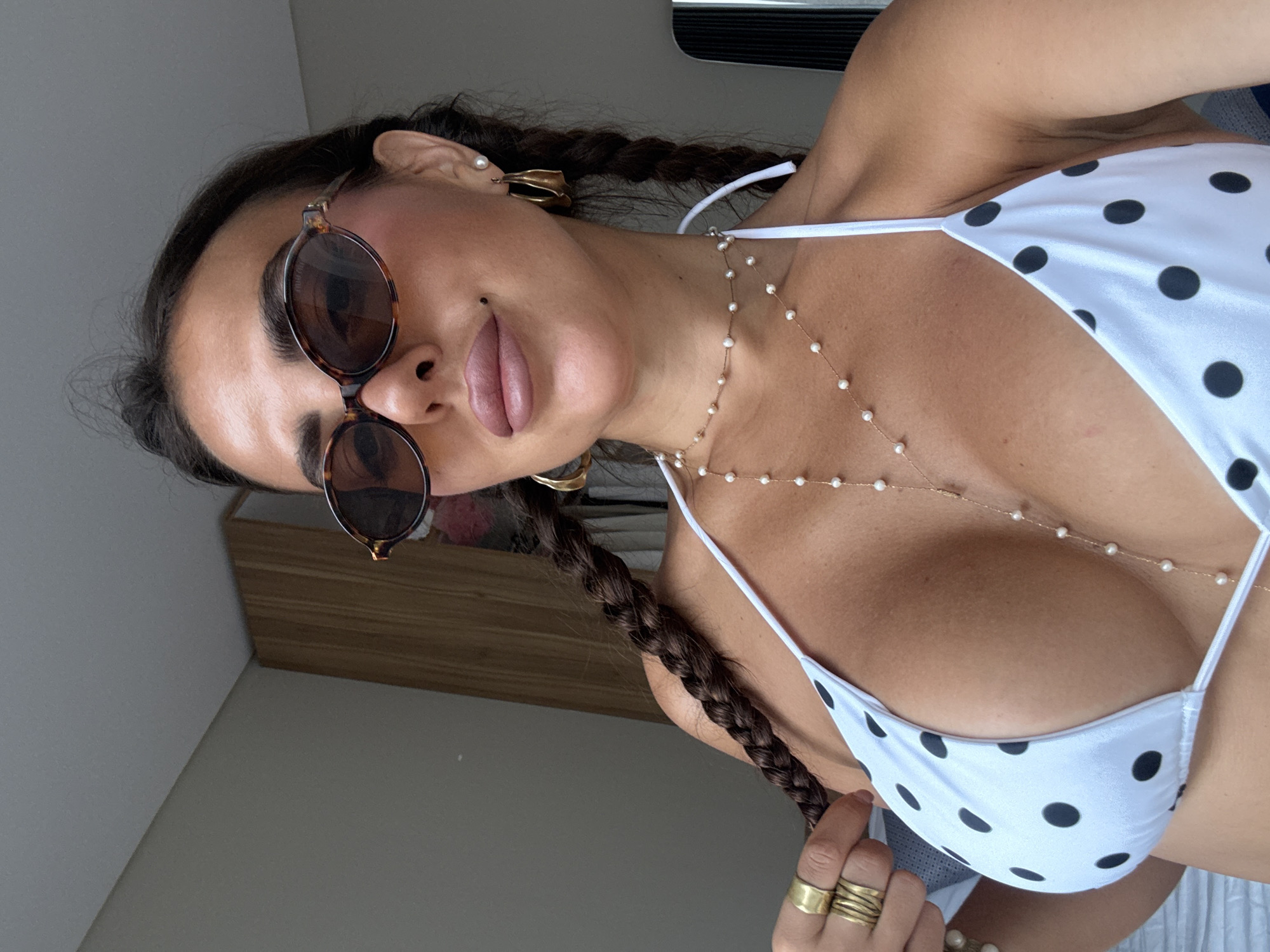 A woman is shown from a low angle in an indoor setting, presumably in Kuta Selatan, Indonesia. She is wearing round, dark sunglasses, gold earrings, and a gold necklace with small pearls that drapes down her chest. She has her hair in braids. She is wearing a white bikini top with black polka dots. She is also wearing several gold rings on her fingers. The background is blurry, but includes a wall, a cabinet and a window.
